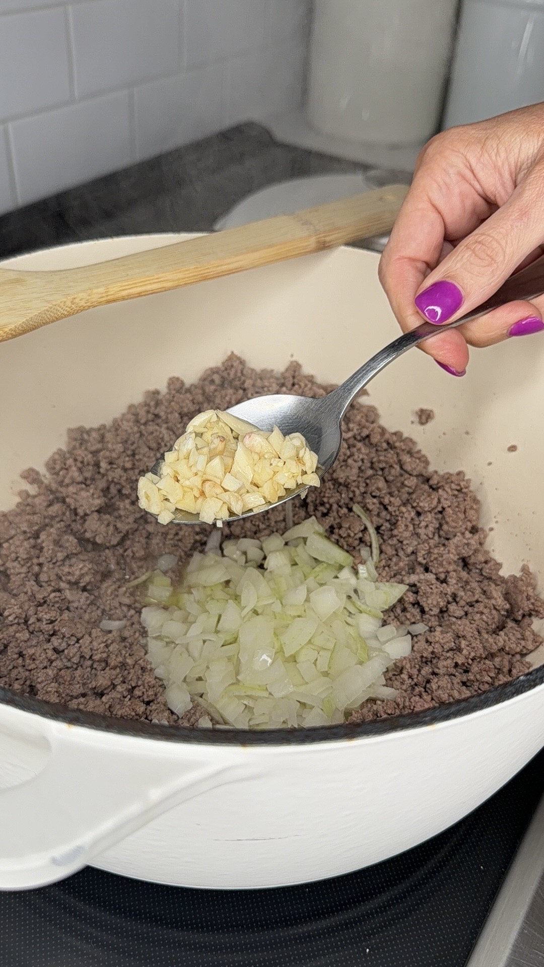 A hand with purple nail polish holds a spoonful of chopped garlic above a pot of sautéed ground beef and diced onions, ready to be added to the mixture.