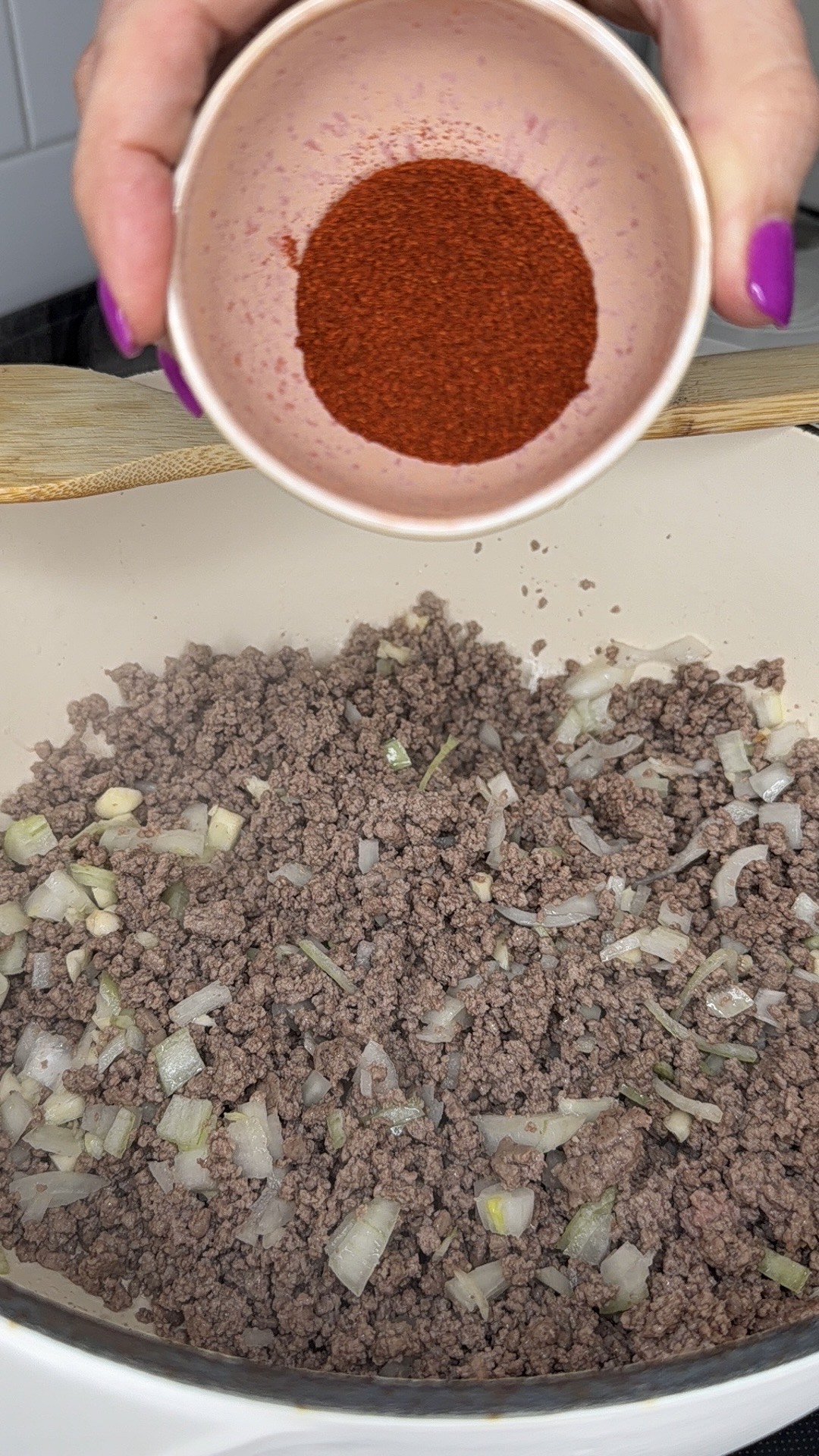 A hand with purple nail polish holds a small bowl of red spice over a pan of browned ground beef and chopped onions.