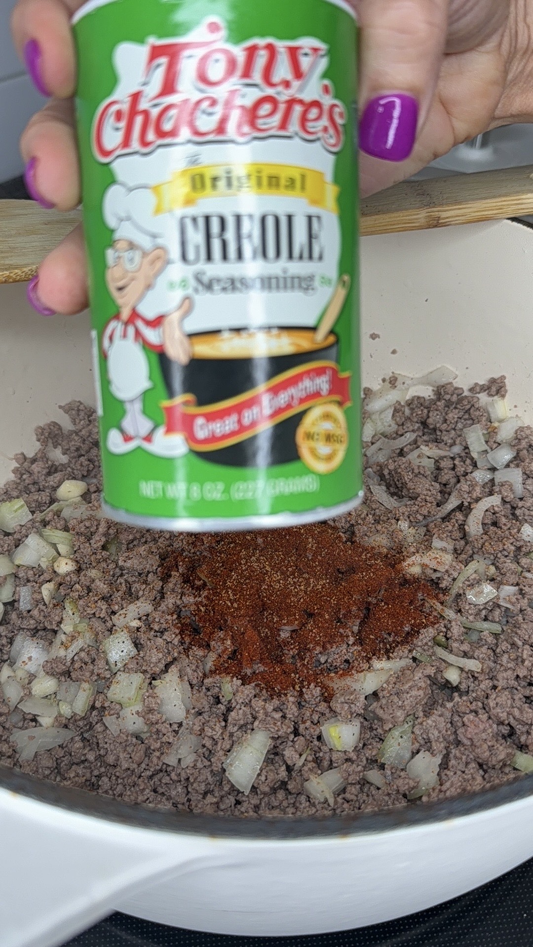 A hand holds a can of Tony Chachere’s Creole Seasoning over a pot of cooked ground beef and diced onions, about to add seasoning. The persons nails are painted purple.