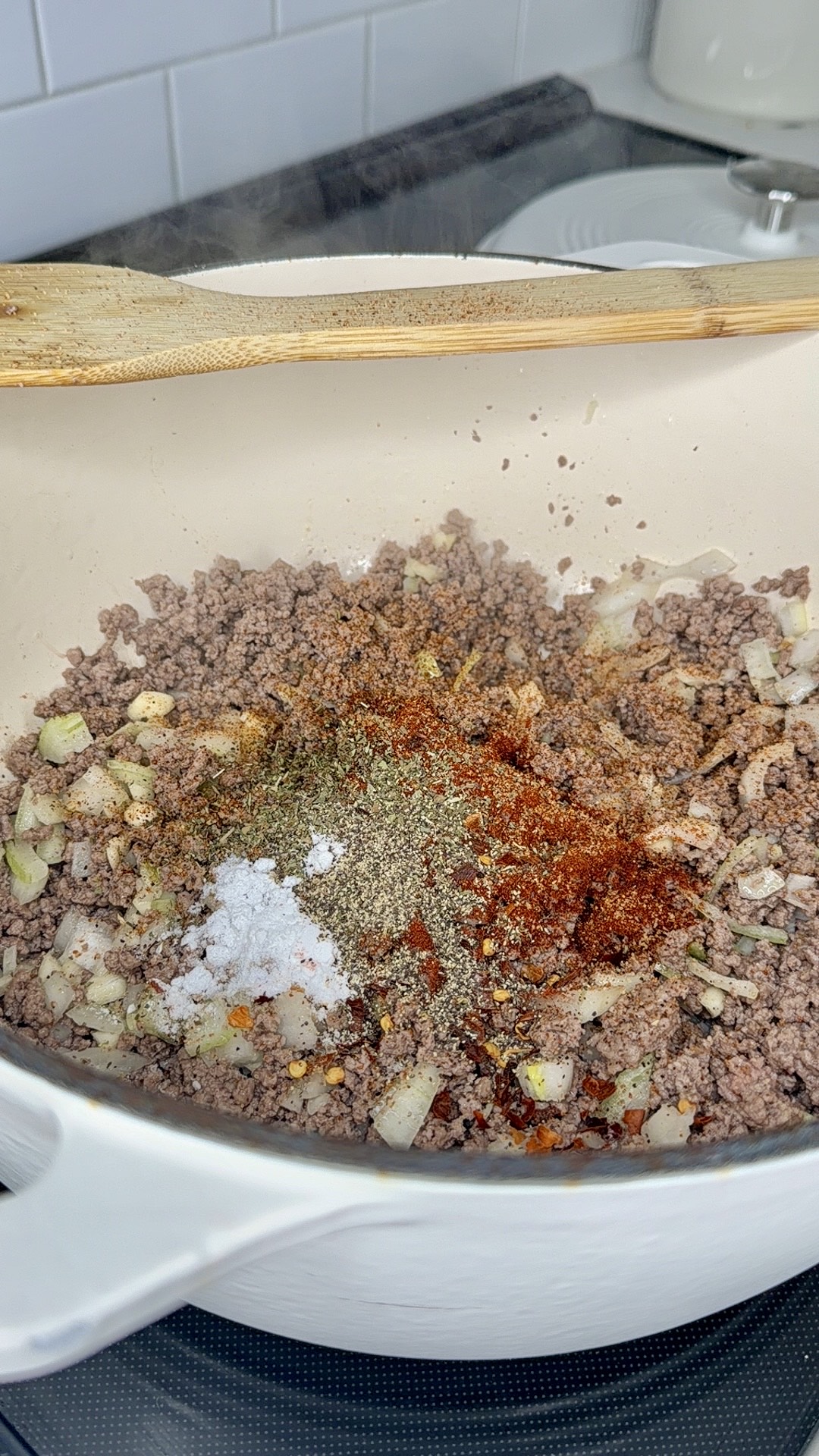 Ground beef and chopped onions are being cooked in a white Dutch oven, with spices including salt, pepper, and paprika sprinkled on top. A wooden spoon rests on the edge of the pot.