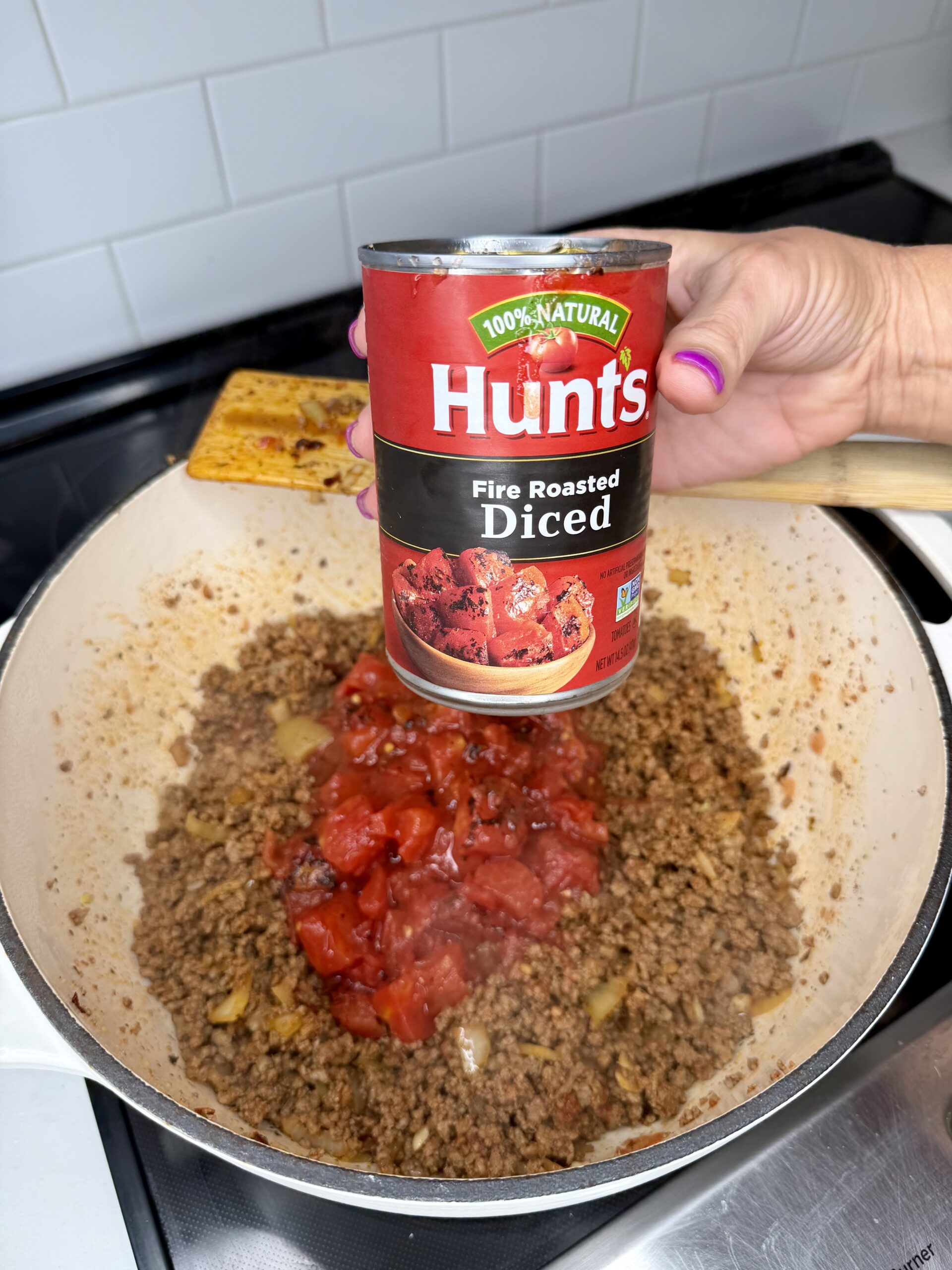 A hand holds a can of Hunts fire roasted diced tomatoes above a pan of browned ground meat and onions on a stovetop, with tomatoes being poured into the pan.