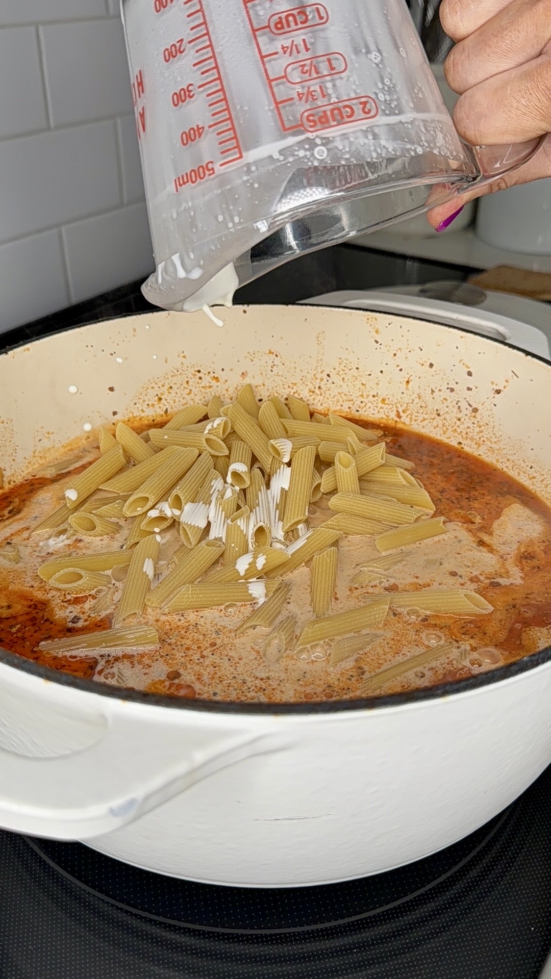 A hand pours water from a measuring cup into a white pot filled with uncooked penne pasta, tomato sauce, and cream on a stove.