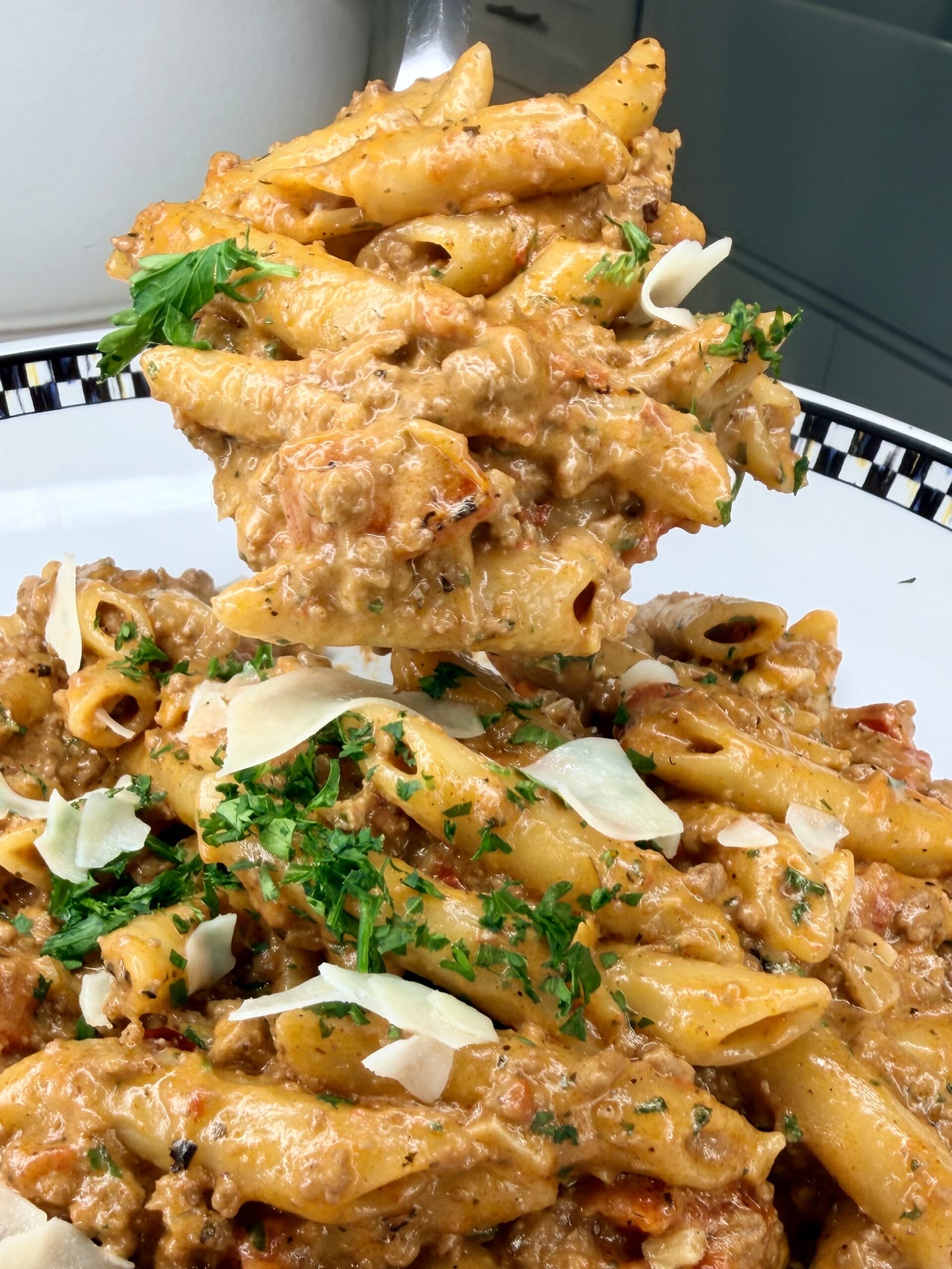 A close-up of creamy penne pasta with ground meat, topped with chopped parsley and shaved Parmesan, being lifted from a white plate with a fork.