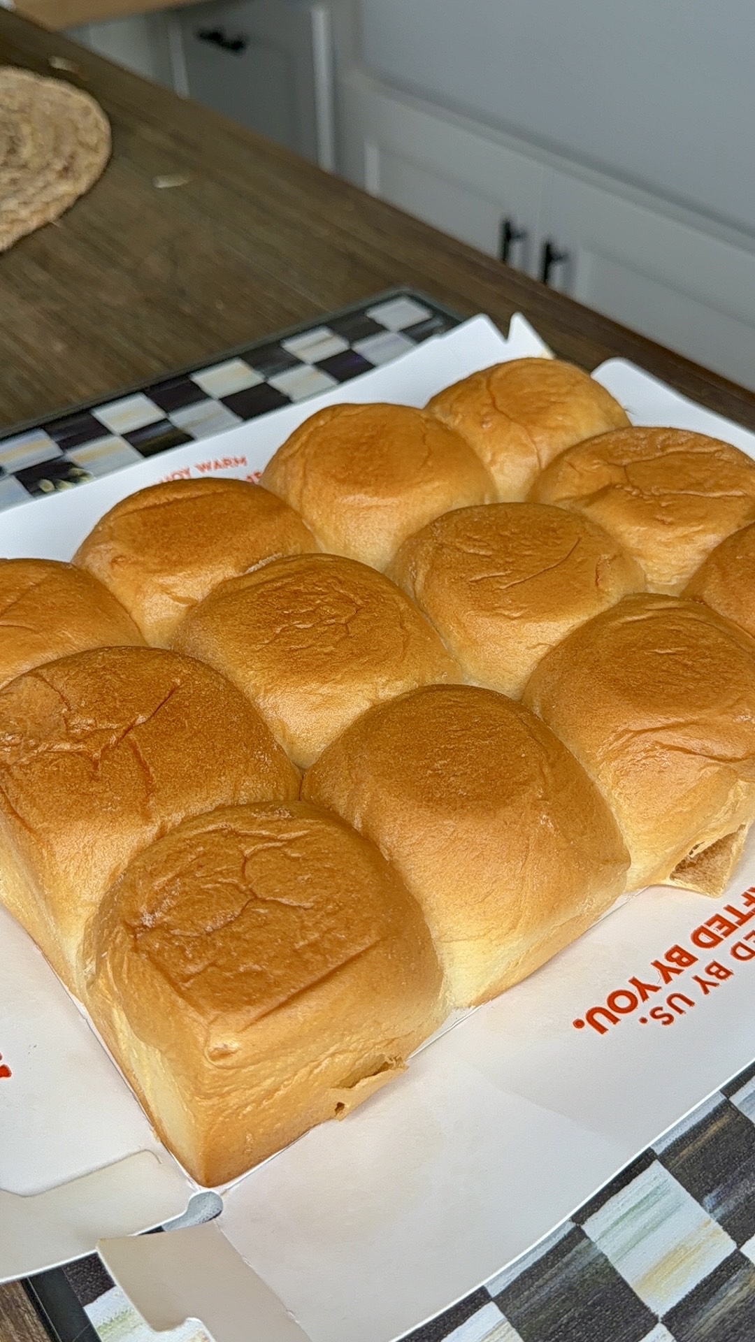 A batch of nine golden-brown dinner rolls sits on a white sheet of paper atop a checkered tray on a kitchen counter. The rolls look soft and freshly baked.
