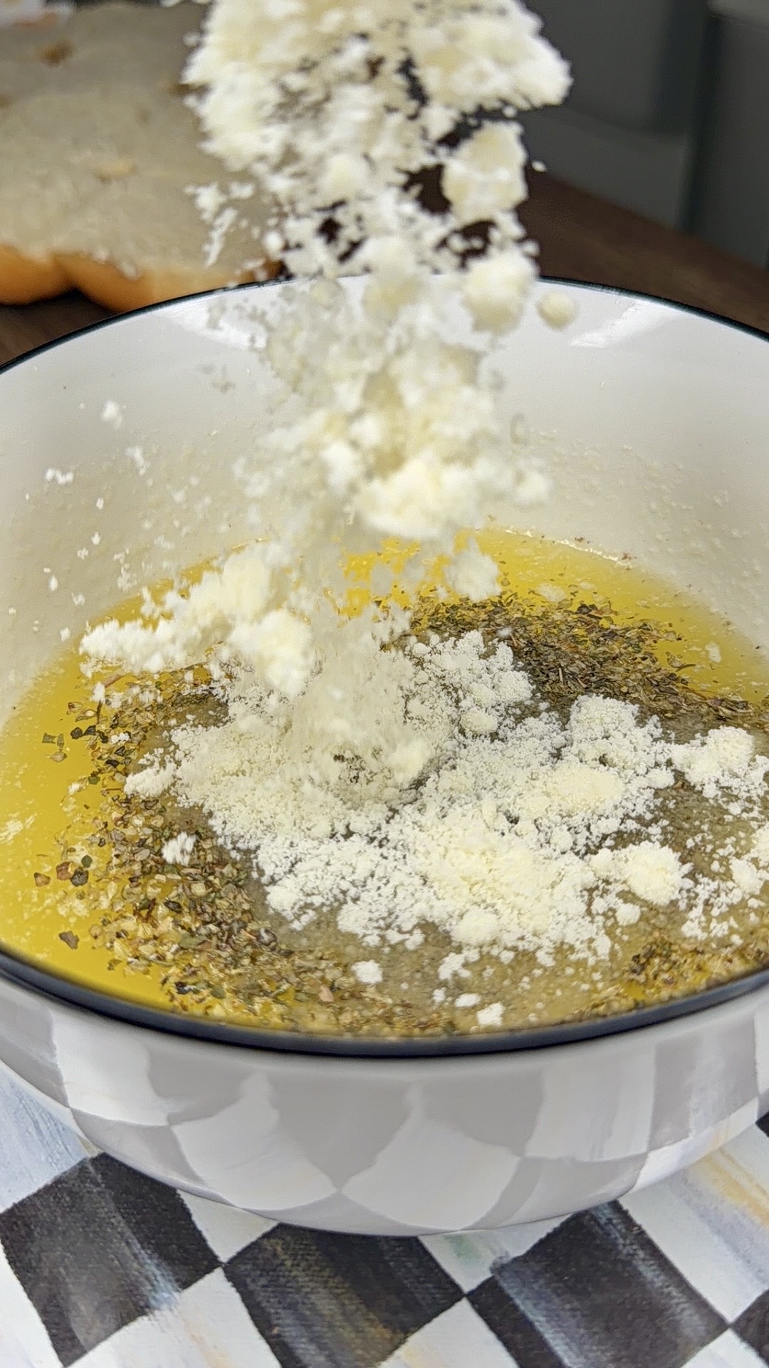 Grated cheese is being sprinkled into a white bowl filled with olive oil and herbs, likely for making a dip or dressing. A piece of bread is visible in the blurred background.