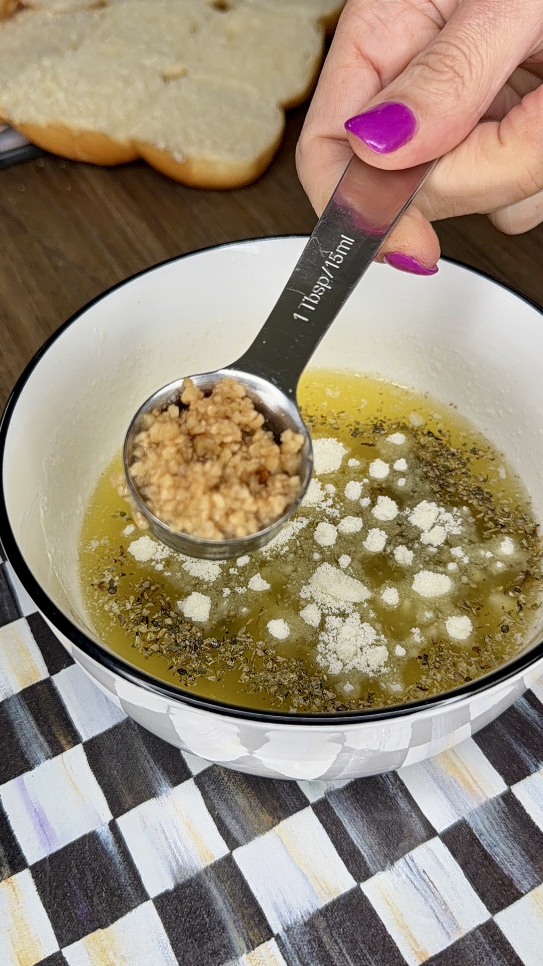 A hand with pink nail polish holds a tablespoon of minced garlic over a bowl containing melted butter, herbs, and grated cheese. Slices of bread are visible in the background.