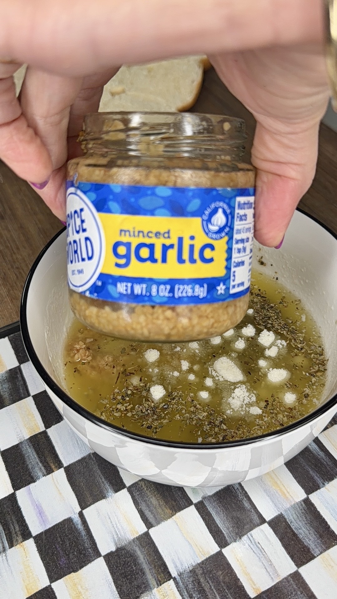 A hand holds a jar of minced garlic above a white bowl filled with olive oil, dried herbs, and seasonings on a checkered placemat. A slice of bread is visible in the background.