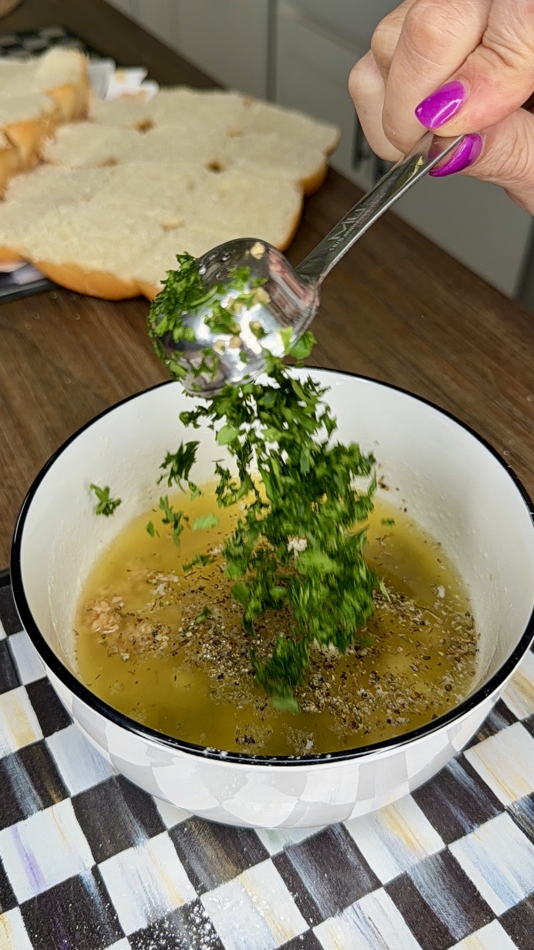 A hand with pink nails adds chopped herbs from a spoon into a bowl of oil, pepper, and spices on a kitchen counter. Slices of bread are visible in the background.