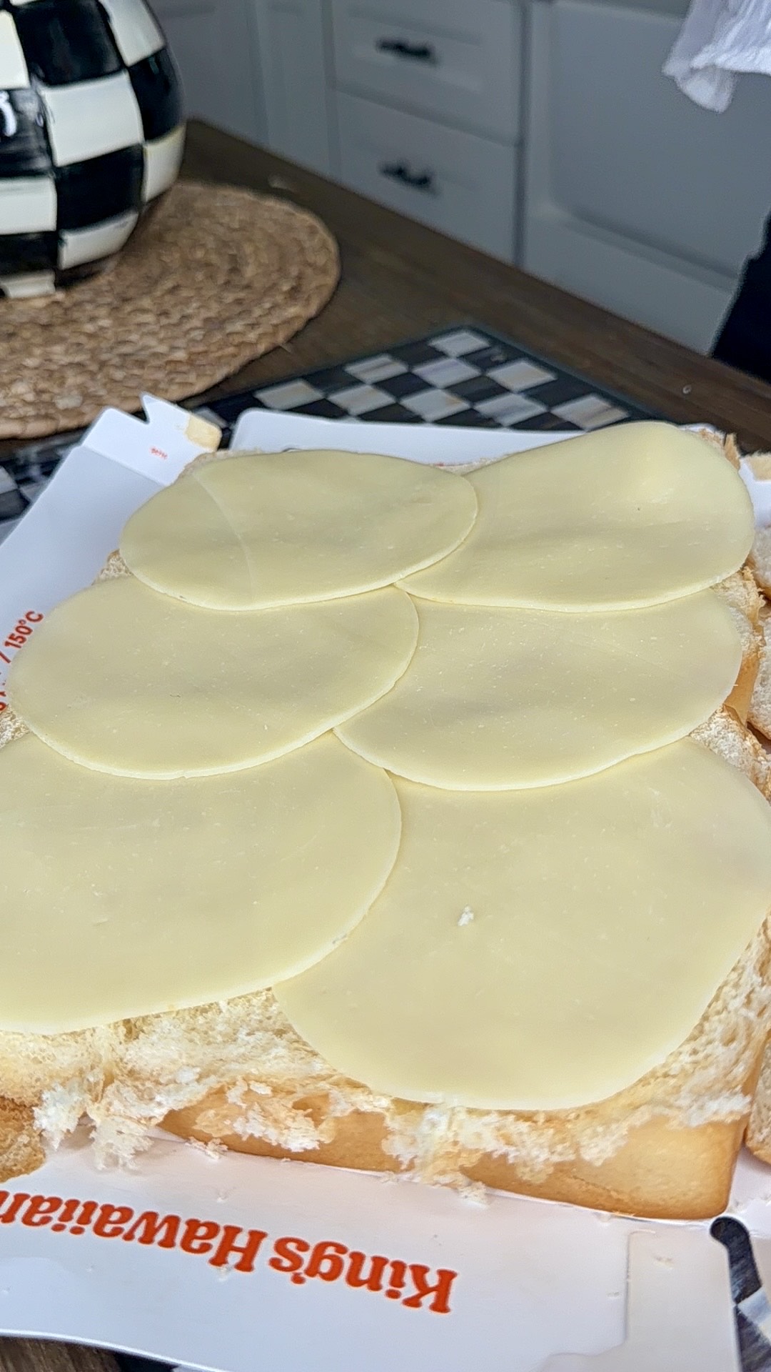 Six slices of provolone cheese are laid out on a piece of white bread covered with mayonnaise, on top of Kings Hawaiian bread packaging, with a kitchen counter and checkered decor in the background.