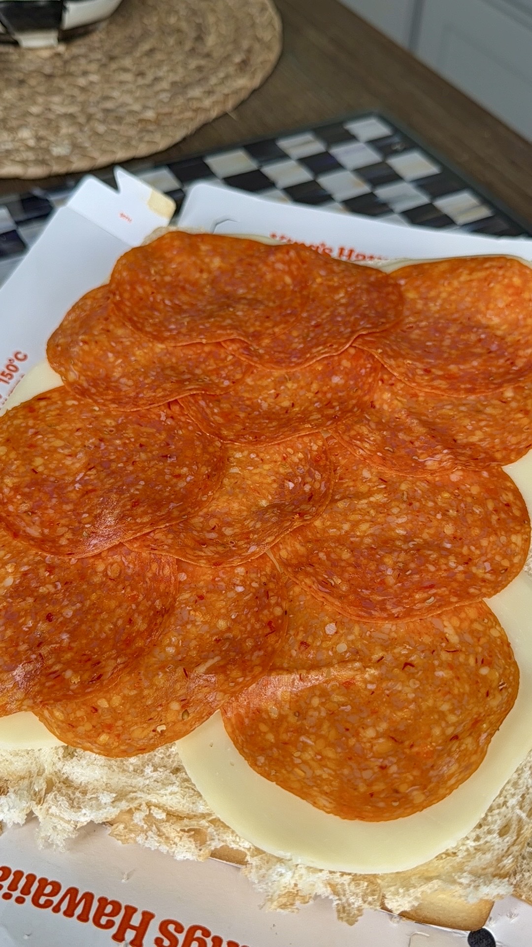 Several slices of pepperoni are layered over white cheese on a sandwich roll, sitting on deli paper, with part of a woven placemat and checkered surface visible in the background.
