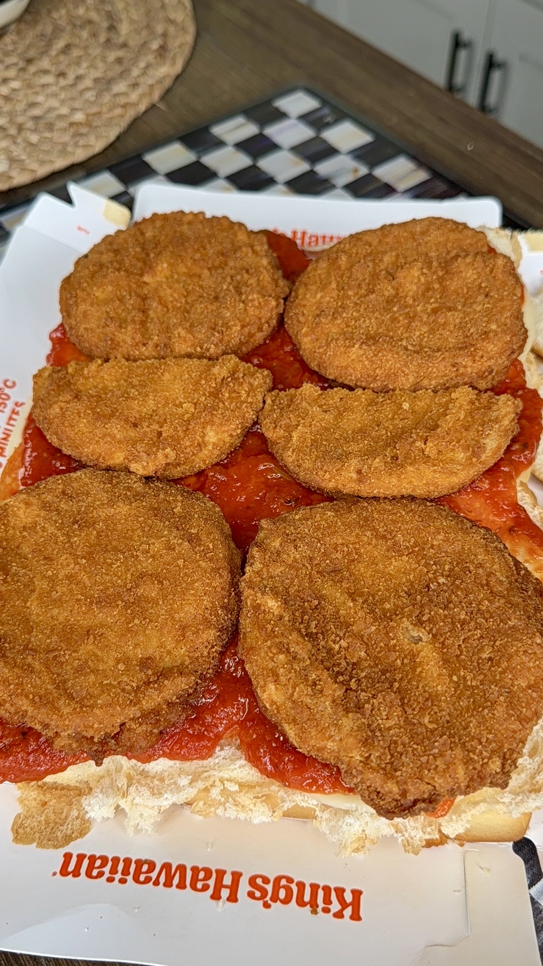 Six breaded and fried eggplant slices are arranged on tomato sauce over a sliced King’s Hawaiian sandwich roll, with a checkered board and woven mat visible in the background.