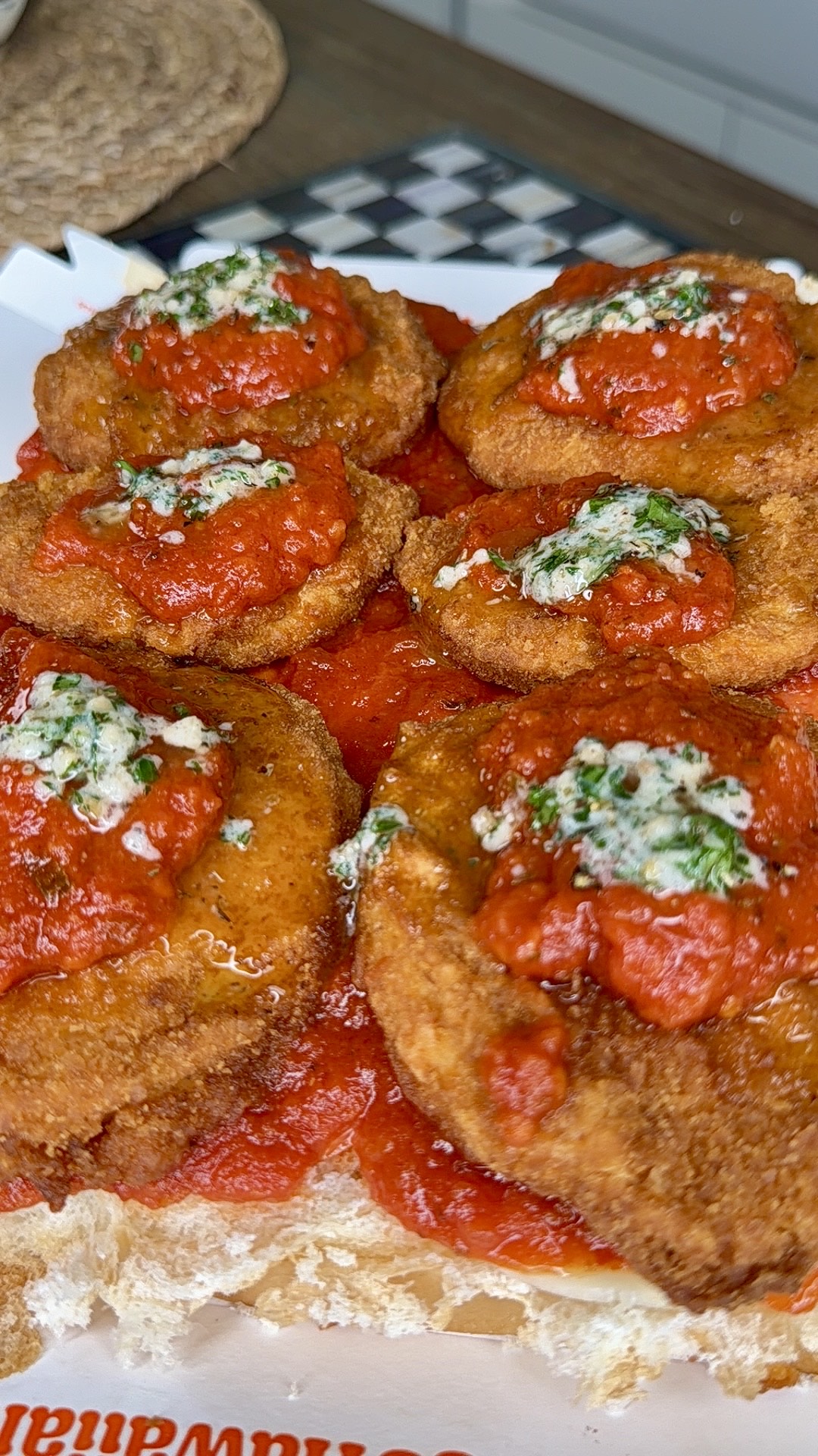 Close-up of several breaded and fried chicken cutlets topped with tomato sauce and a sprinkle of green herbs, served on a sandwich roll. The food looks crispy and saucy, ready to eat.