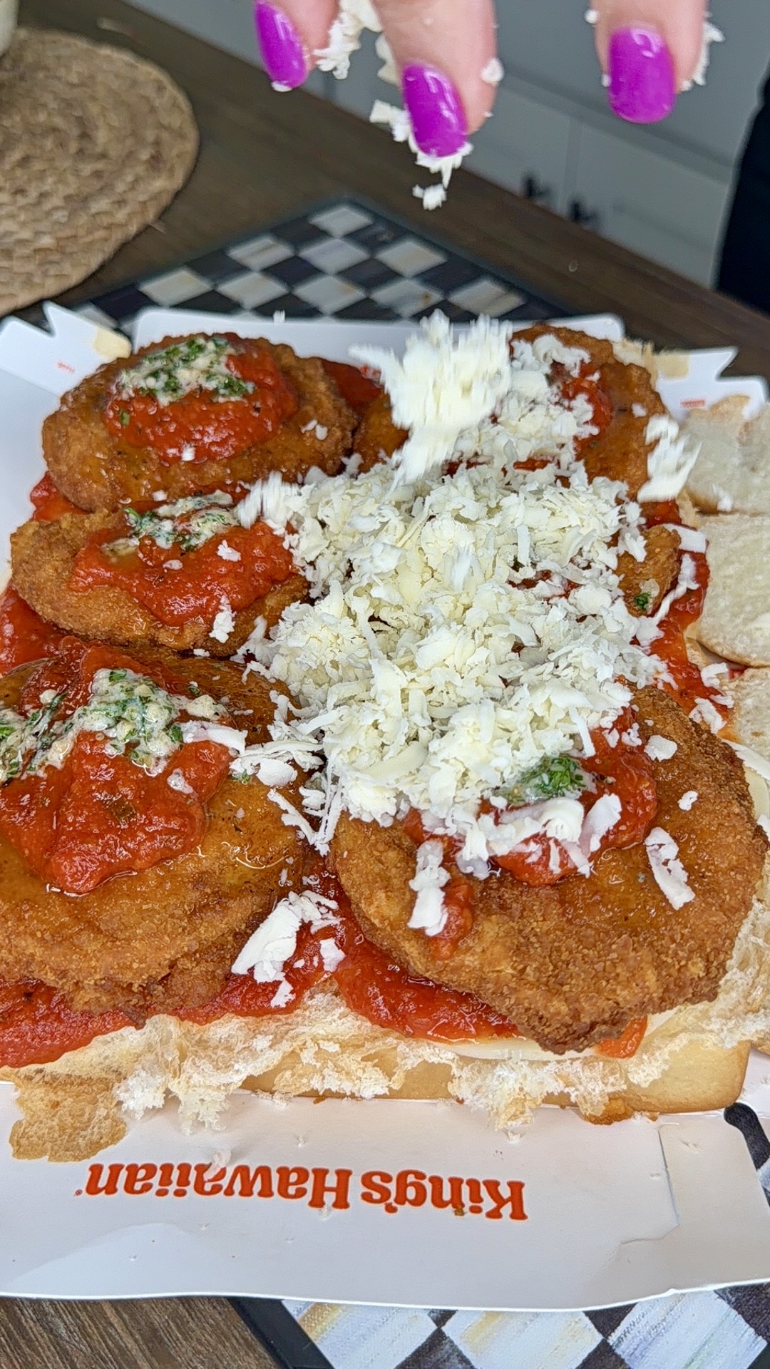 A hand with bright purple nails sprinkles shredded cheese onto a sandwich filled with breaded and fried cutlets topped with marinara sauce, all placed on a hoagie roll on a Kings Hawaiian wrapper.