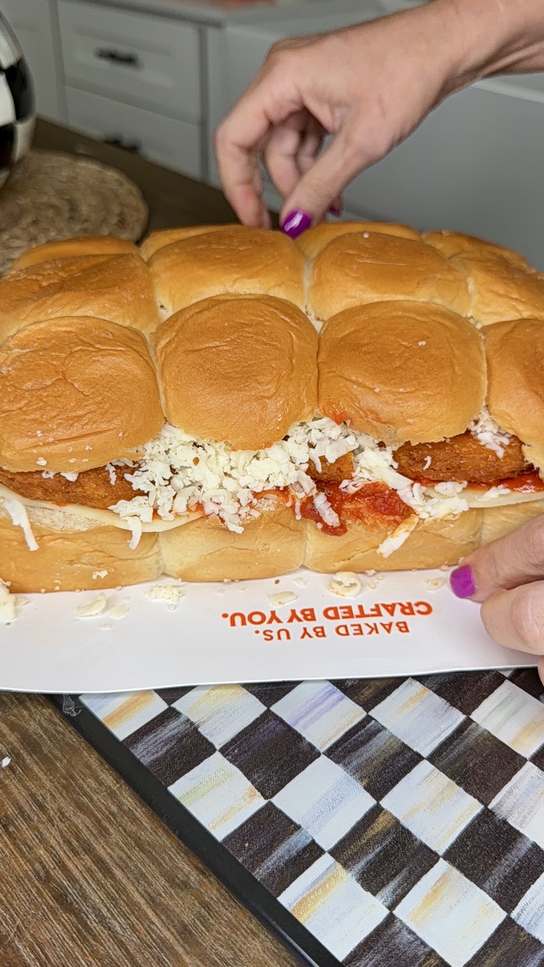 A person with purple nail polish slices a sandwich made of bread rolls filled with breaded chicken, marinara sauce, and shredded cheese on a kitchen counter.