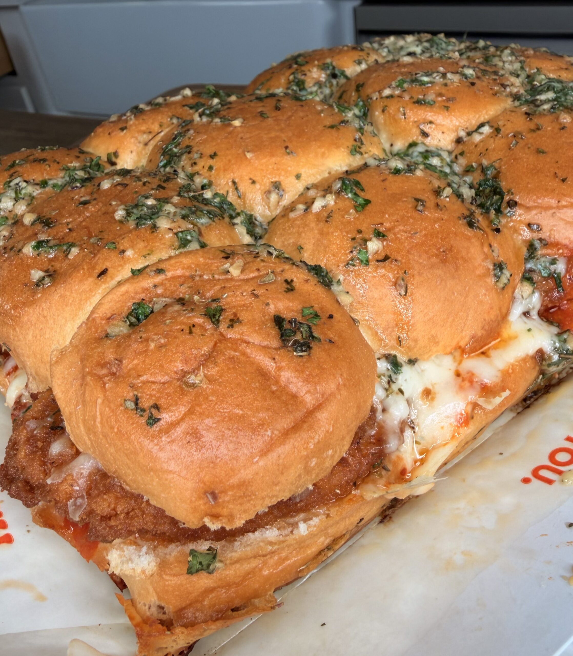A close-up of a tray of cheesy chicken sliders on soft, golden buns topped with herbs and garlic, with melted cheese and sauce visible between the layers.