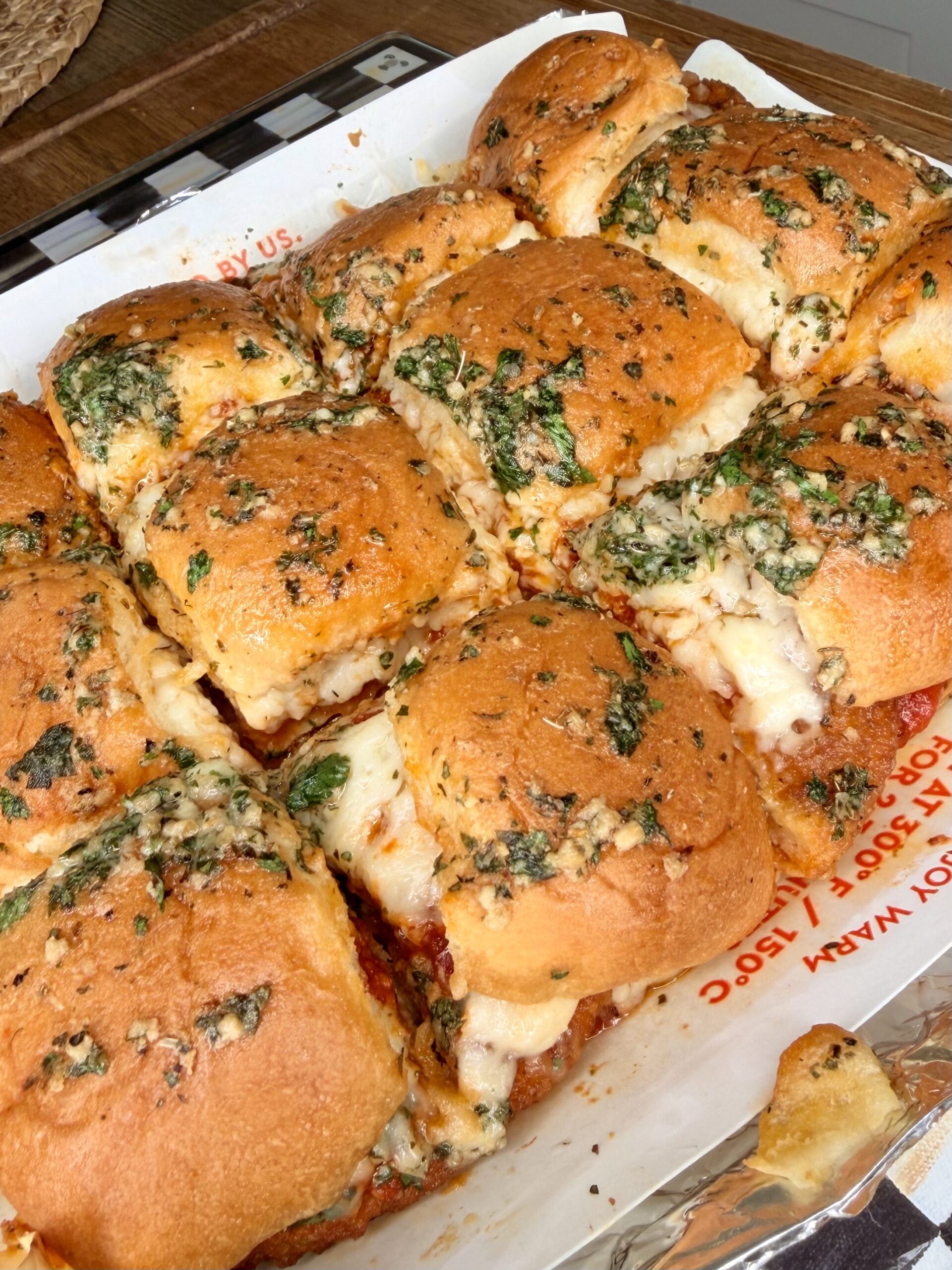 Golden, cheesy baked sliders topped with melted cheese, garlic, and herbs sit on a foil-lined tray. The sliders are closely packed, with gooey cheese oozing from between the soft, browned buns.