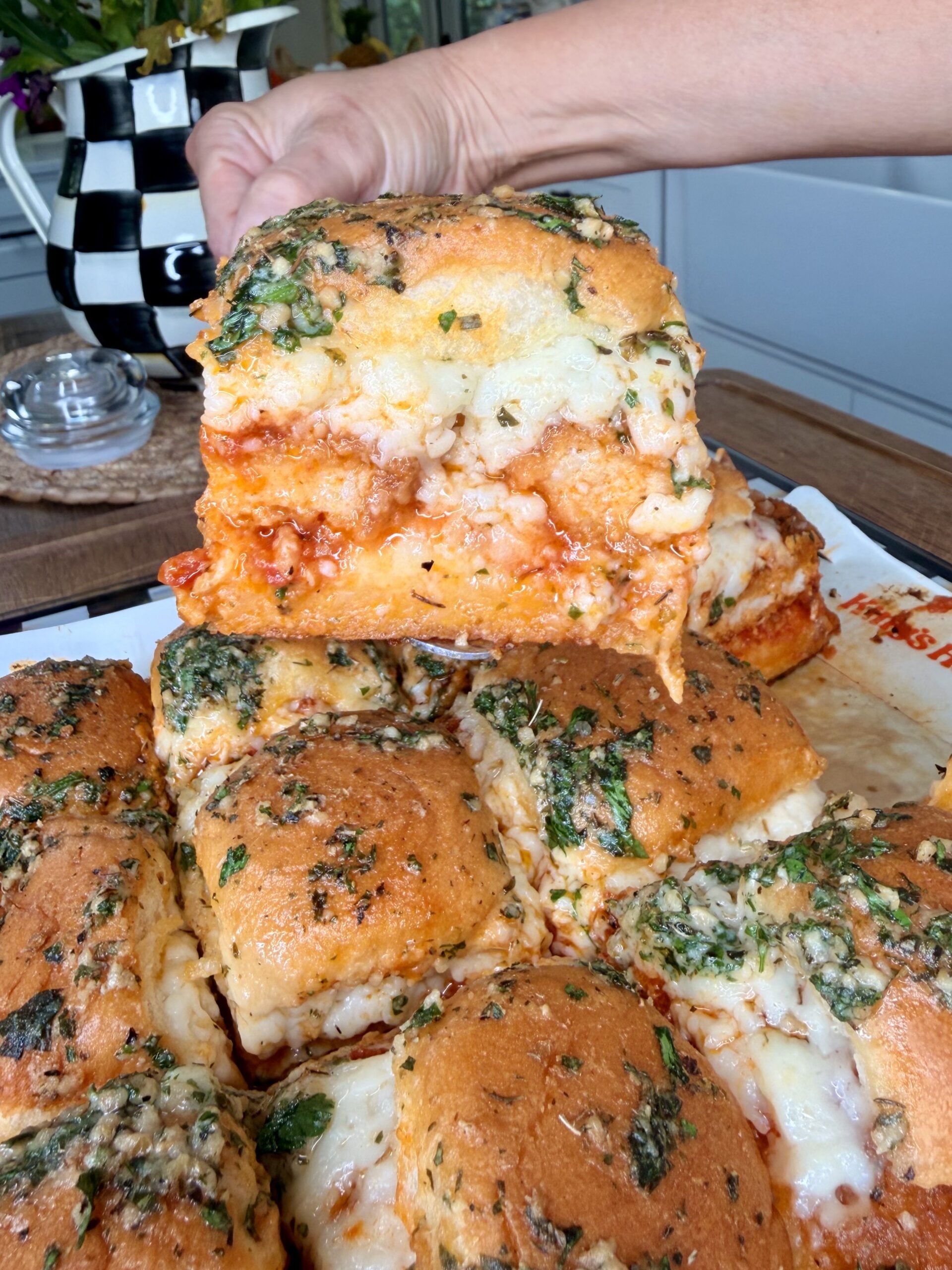 A hand lifts a cheesy, herb-topped baked sandwich filled with tomato sauce and melted cheese from a tray of similar sandwiches in a kitchen setting.