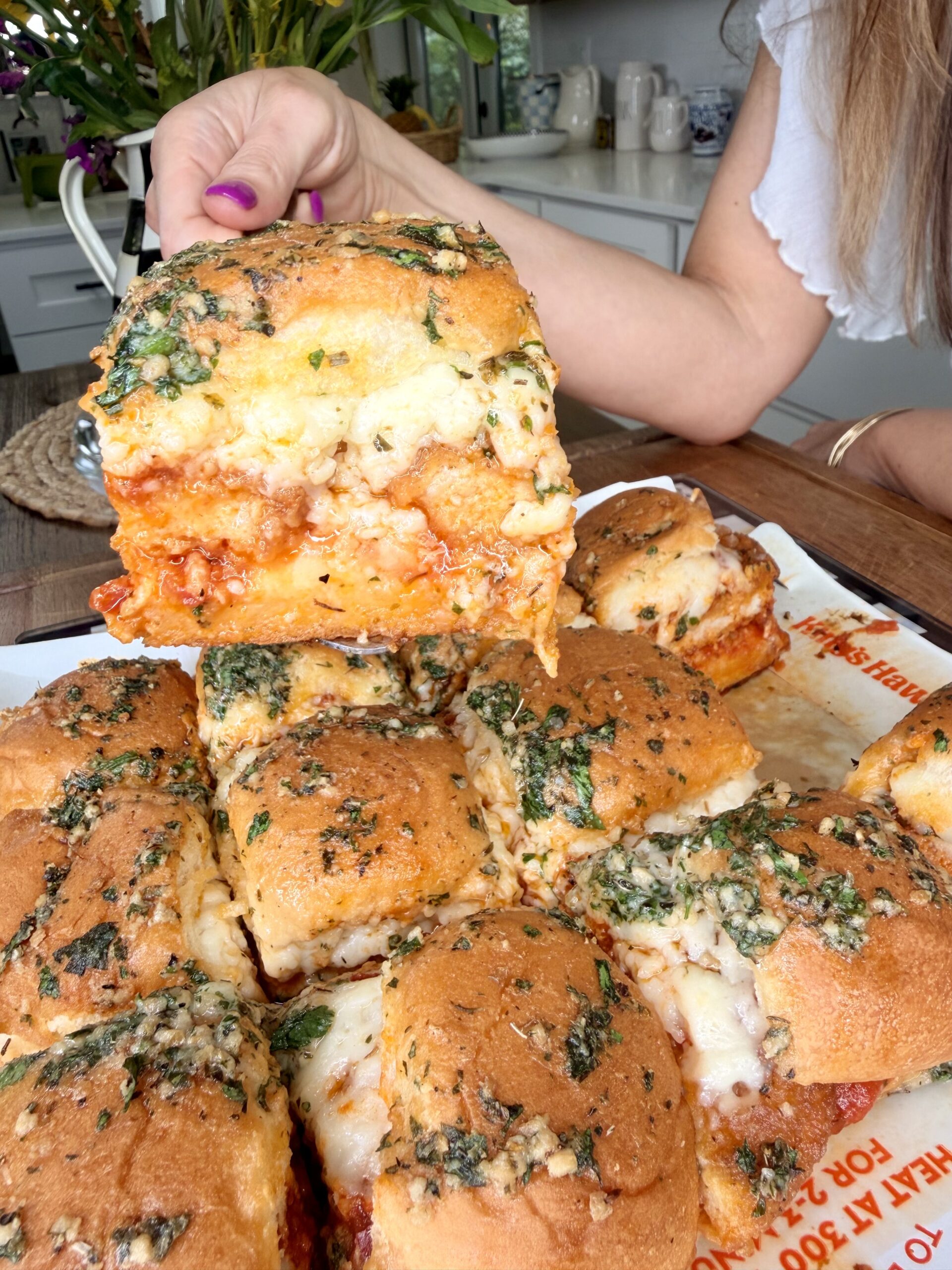 A hand lifts a cheesy, herb-topped sandwich from a tray of baked sliders filled with marinara sauce and melted cheese in a kitchen setting.