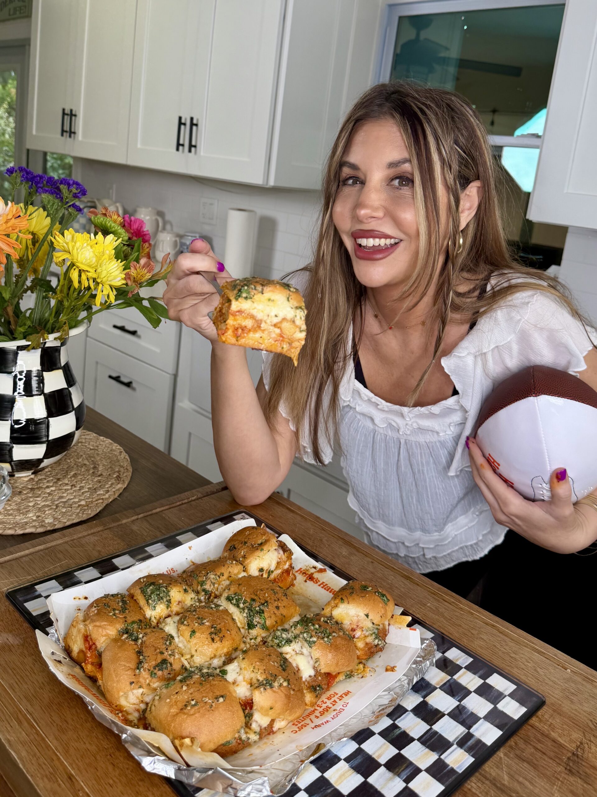 A smiling woman holding a football in one hand and a cheesy garlic bread roll in the other, standing in a kitchen next to a tray of bread rolls and a vase of colorful flowers.