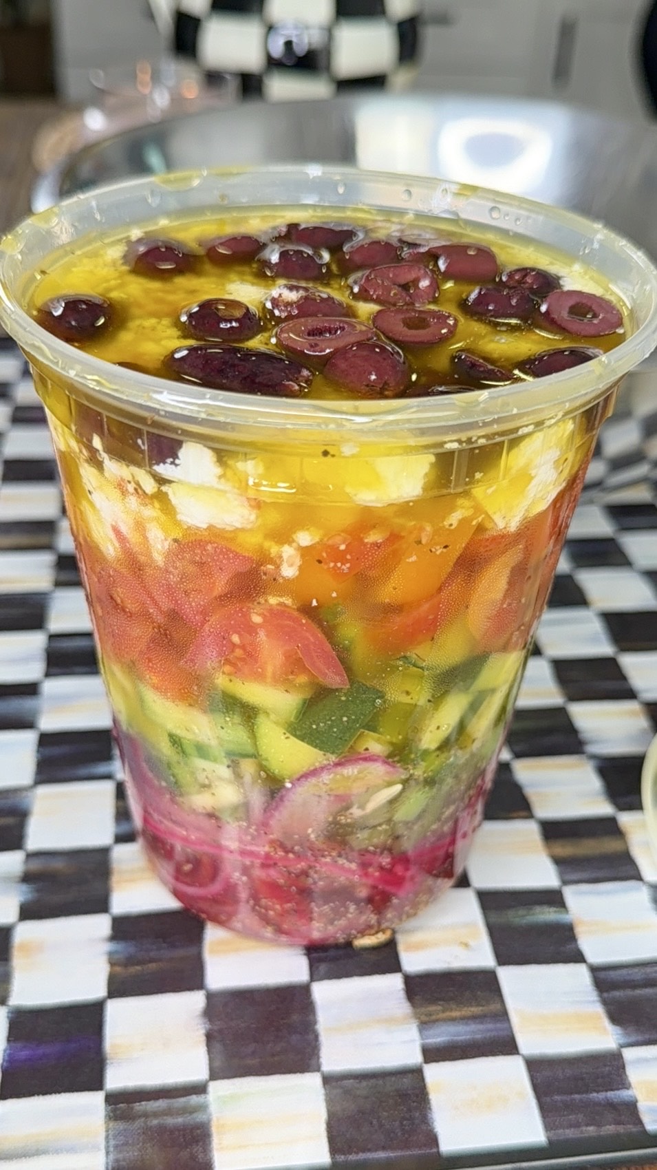 A clear plastic cup filled with layered salad, including chopped onions, cucumbers, tomatoes, feta cheese, olives, and topped with olive oil, sits on a checkered table surface in a kitchen setting.