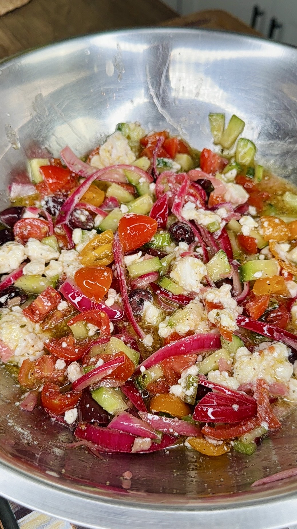 A stainless steel bowl filled with a colorful salad of chopped tomatoes, red onions, cucumbers, olives, and crumbled feta cheese, mixed with a dressing.