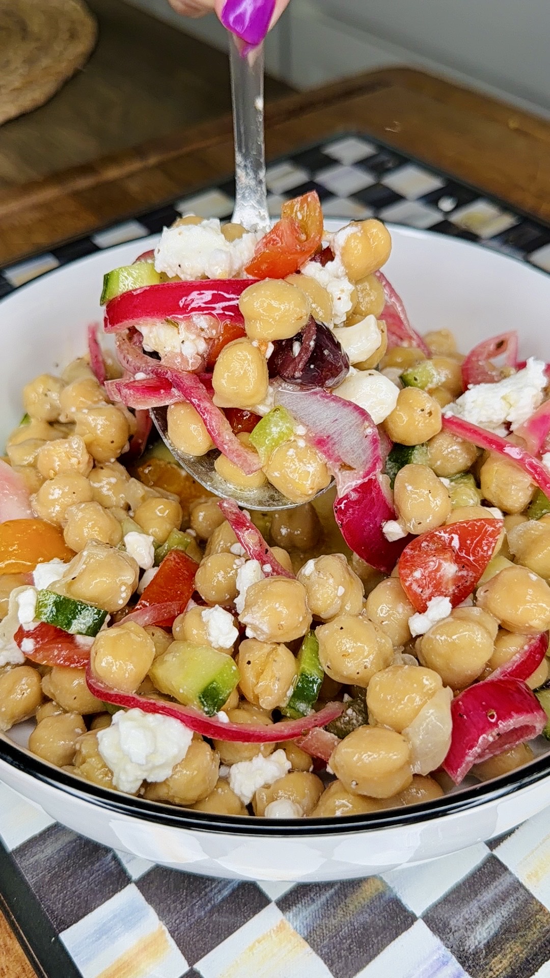 A bowl of chickpea salad with chopped tomatoes, cucumbers, red onions, olives, and crumbled feta cheese. A spoon is lifting a portion, showcasing the colorful, fresh ingredients.