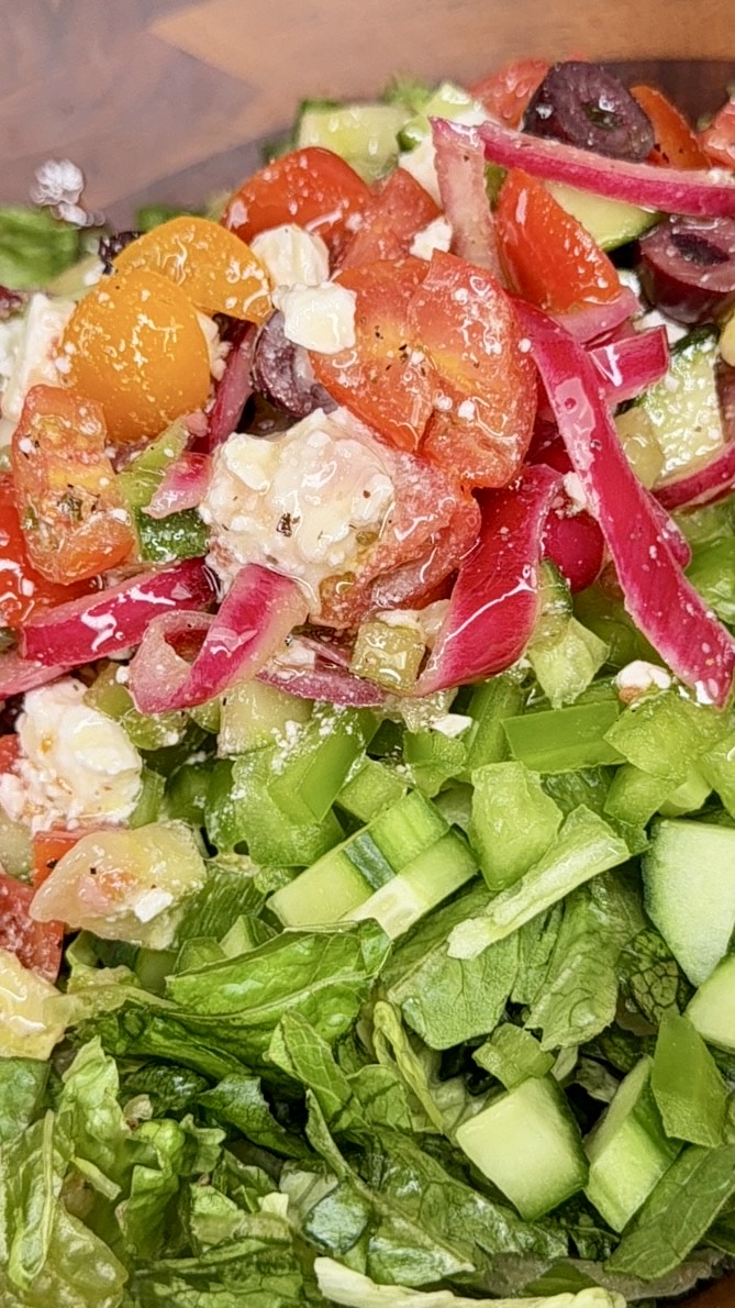 A close-up of a colorful salad featuring chopped lettuce, cucumbers, tomatoes, red onions, olives, feta cheese, and possibly bell peppers, all lightly dressed and mixed together.