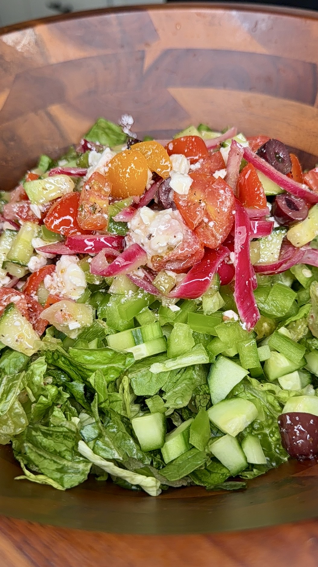 A wooden bowl filled with a colorful salad, including chopped romaine lettuce, diced cucumbers, grape tomatoes, red onions, olives, pepperoncini, and crumbled feta cheese.