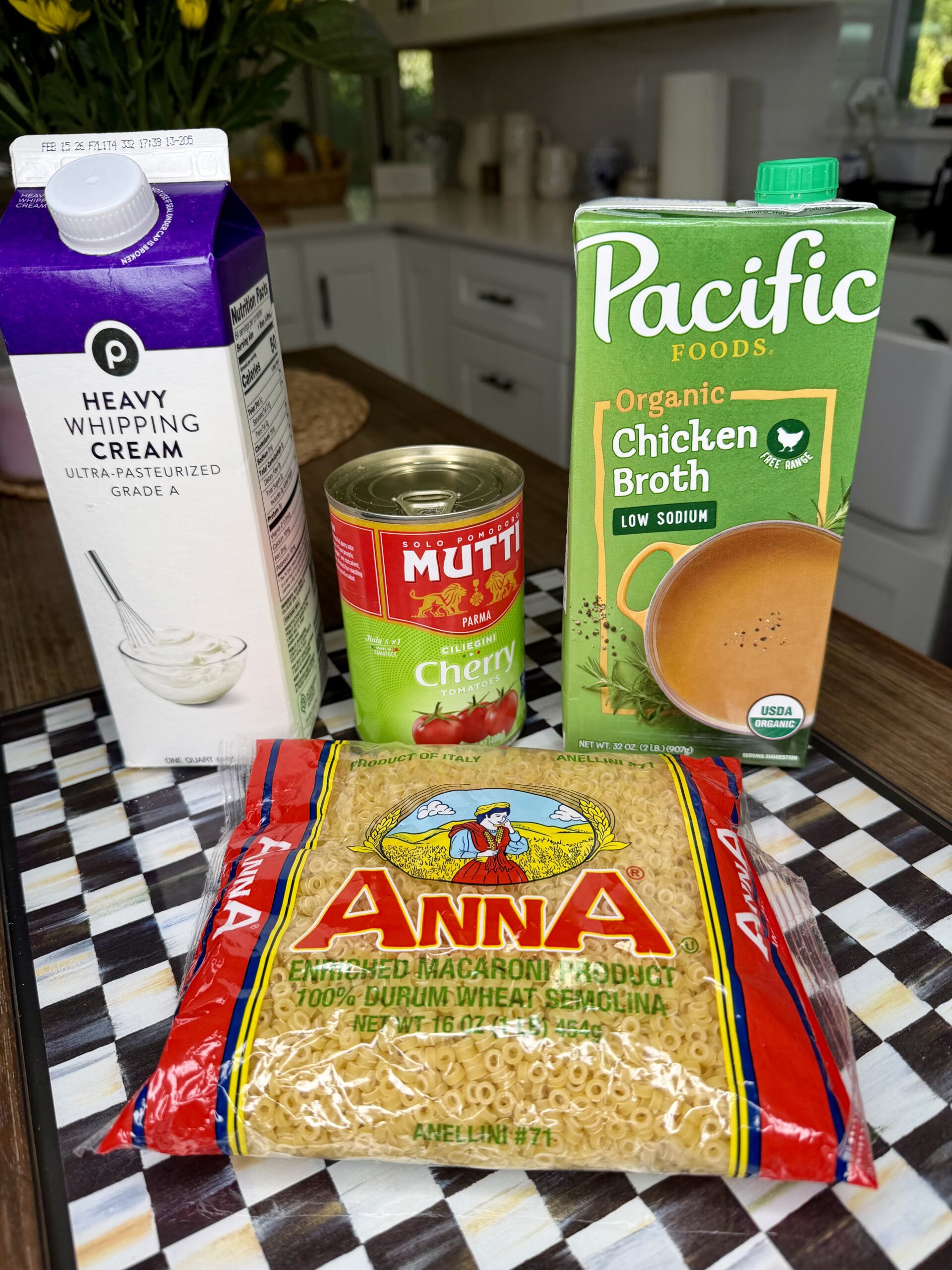A carton of heavy whipping cream, a carton of Pacific Foods organic chicken broth, a can of Mutti cherry tomatoes, and a bag of Anna ditalini pasta are arranged on a checkered countertop in a kitchen.