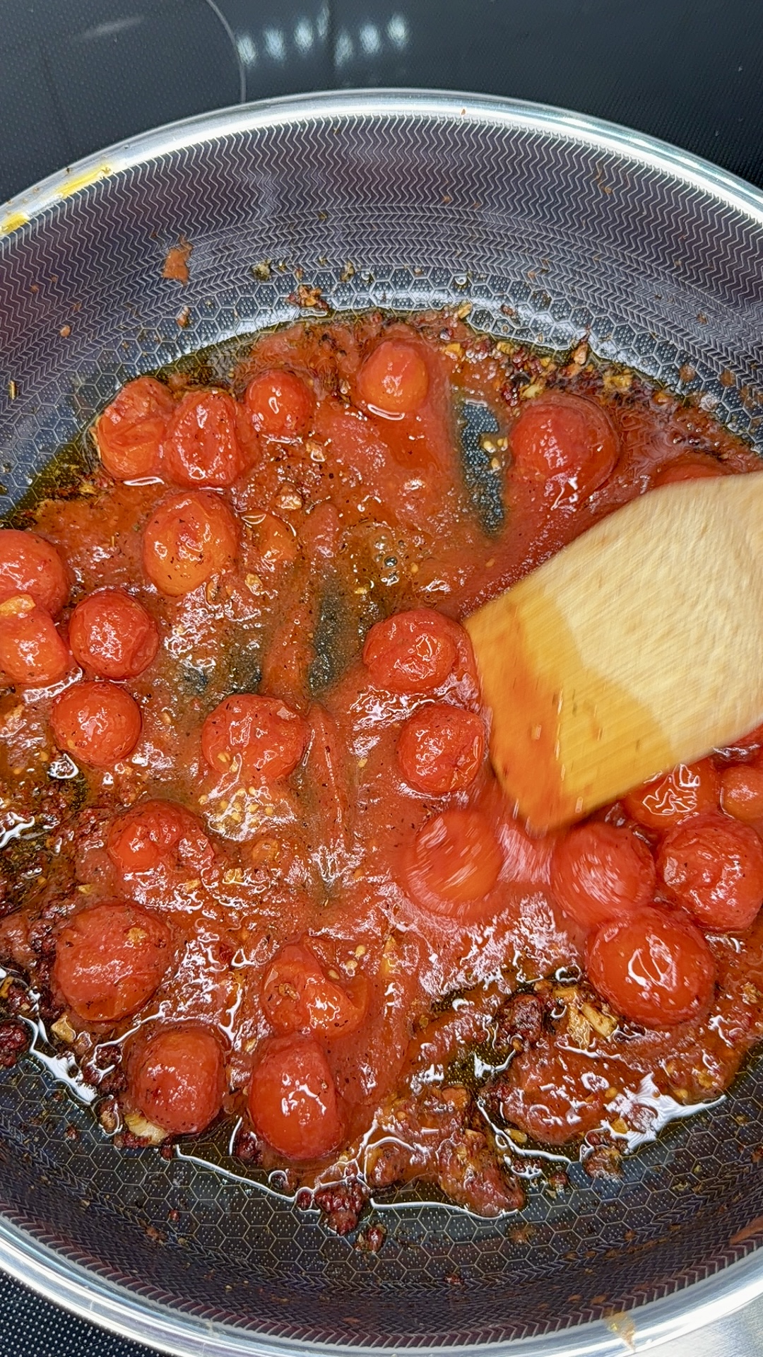 Cherry tomatoes cooking in a nonstick pan with oil and spices, being stirred with a wooden spatula; the tomatoes are beginning to break down and release their juices.