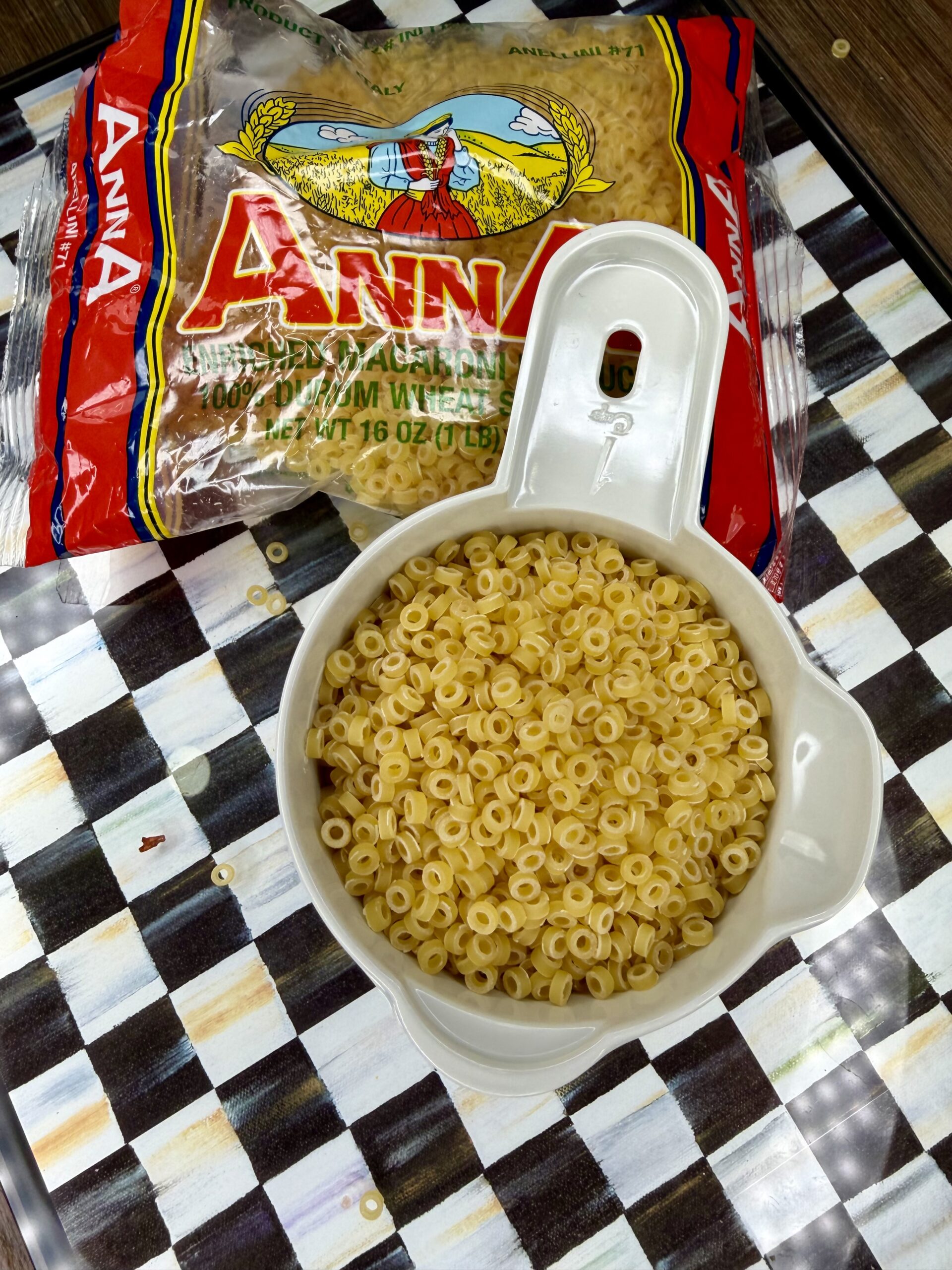 A measuring cup filled with dry ditalini pasta sits on a checkered surface, with a partially opened bag of Anna brand ditalini pasta in the background. Some pasta pieces are scattered around.