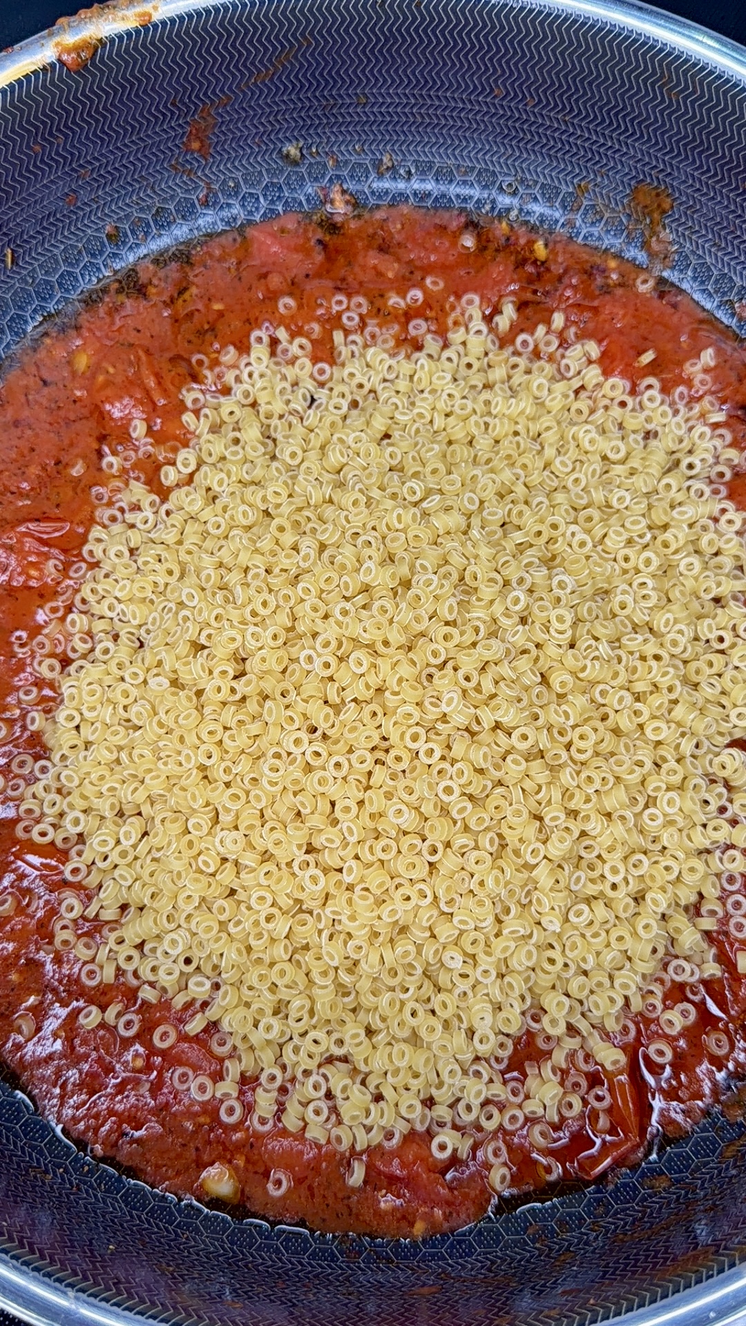 A close-up of a pan with tomato sauce and a large pile of dry ring-shaped pasta sitting on top, not yet mixed together.