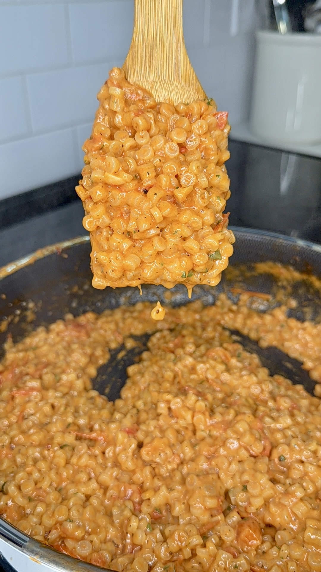 A wooden spoon holds up a serving of creamy, saucy pasta over a pan filled with the same pasta. The sauce appears orange, likely tomato-based, and the pasta is small and round. The background shows a kitchen counter.