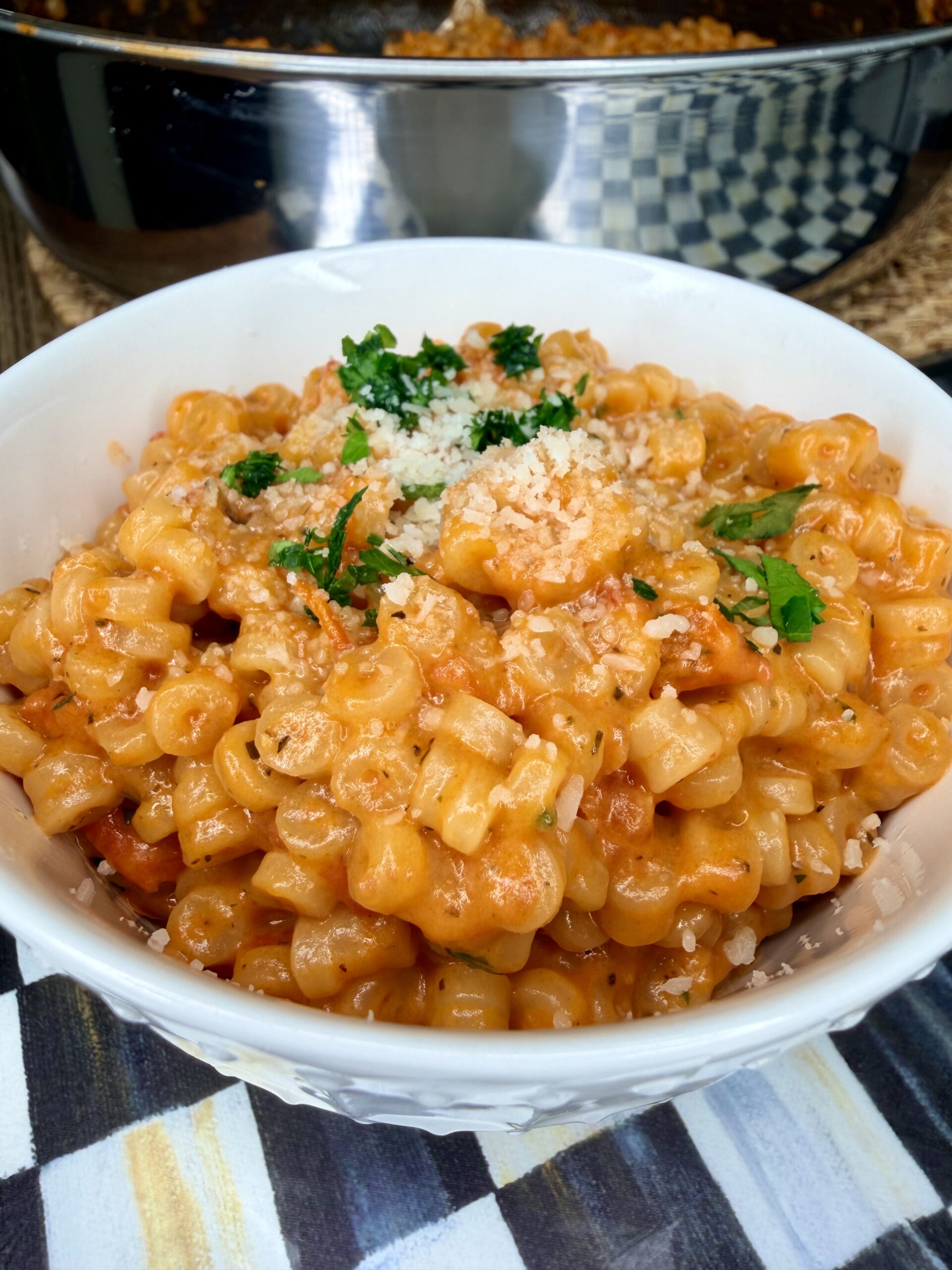 A bowl of creamy pasta with small tube-shaped noodles in a rich orange sauce, topped with grated cheese and fresh parsley. A pan of more pasta is visible in the background.