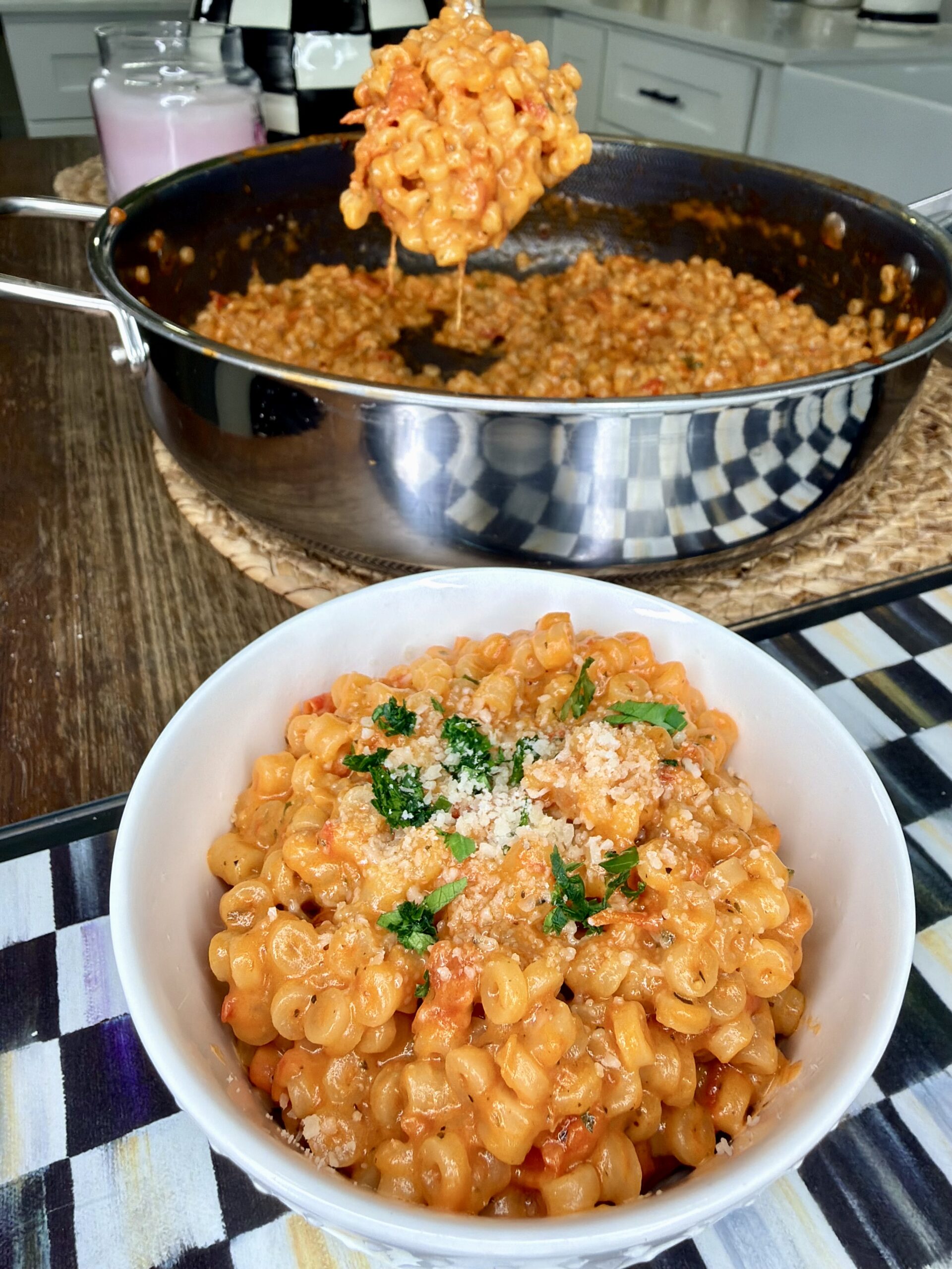 A bowl of creamy macaroni pasta topped with herbs and breadcrumbs sits on a table, with a large pan of the same pasta in the background and a serving spoon lifting a portion from the pan.