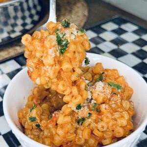 A hand with purple nail polish holds a spoonful of pasta in red sauce, garnished with herbs and grated cheese, over a white bowl on a checkered table mat.