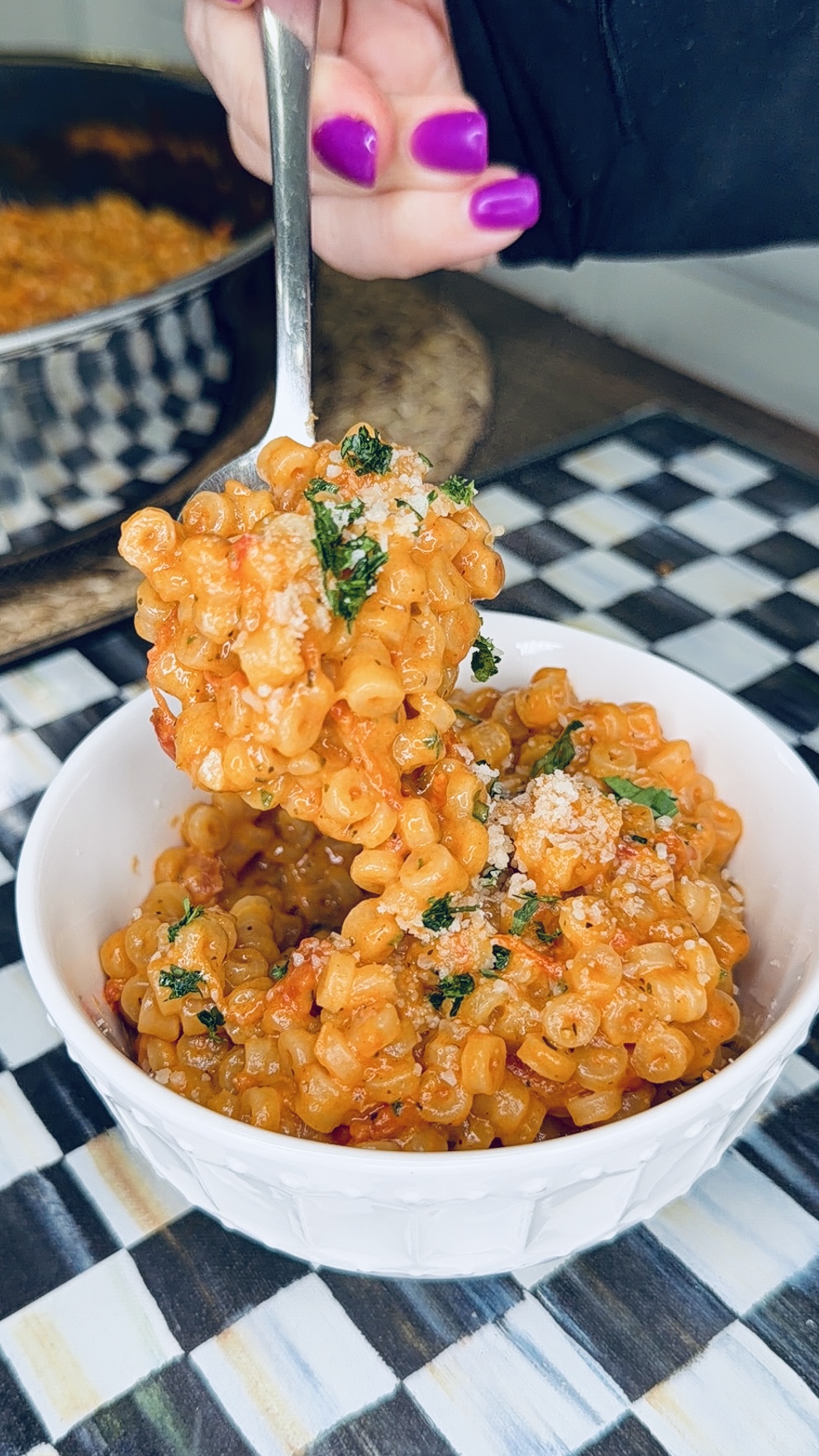 A hand with purple-painted nails holds a spoonful of pasta in red sauce, topped with herbs and grated cheese, over a white bowl on a checkered surface.