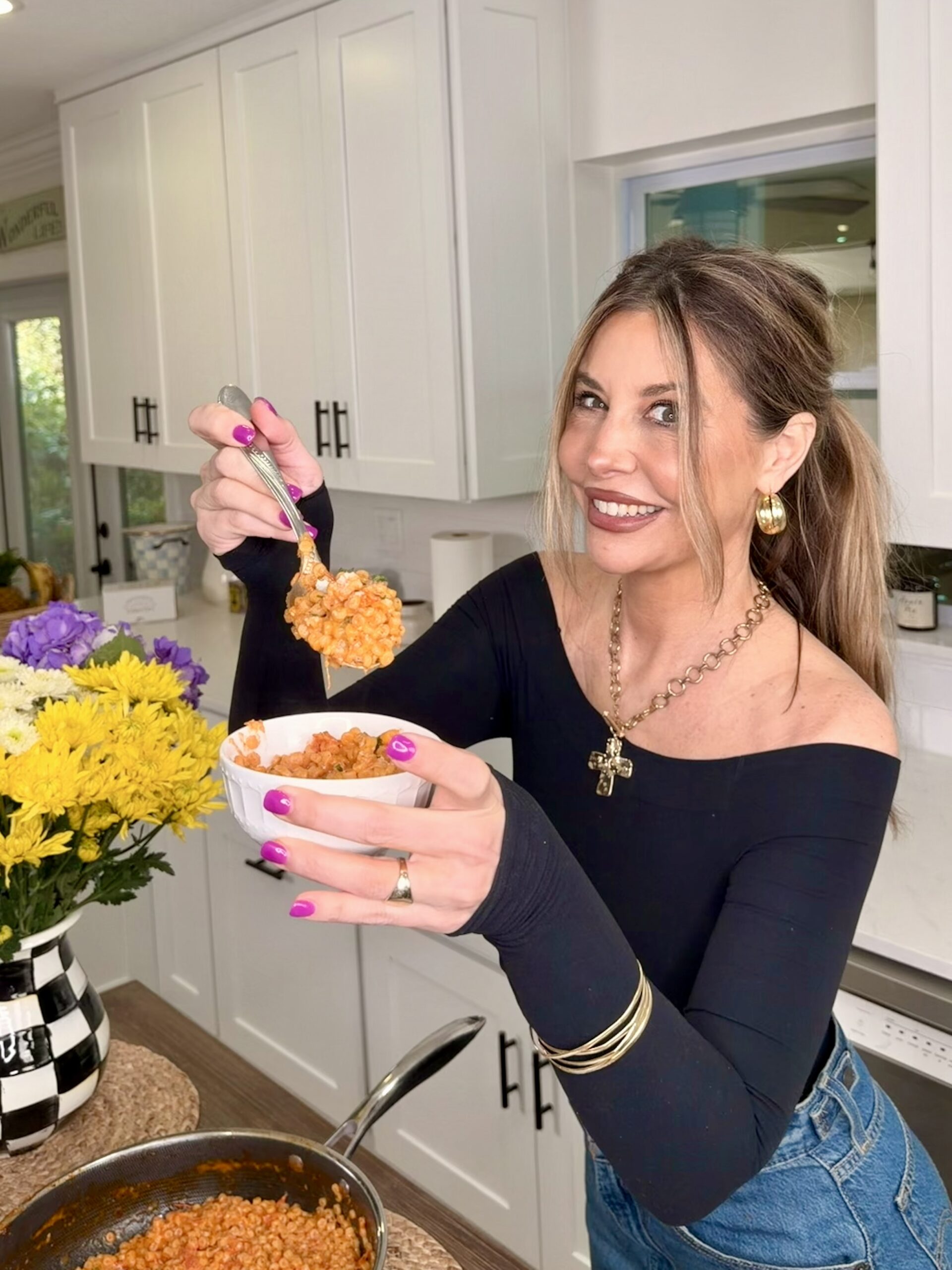 A smiling woman with long brown hair holds a bowl of food in one hand and a spoonful in the other, standing in a bright kitchen with white cabinets and a vase of yellow and purple flowers on the counter.