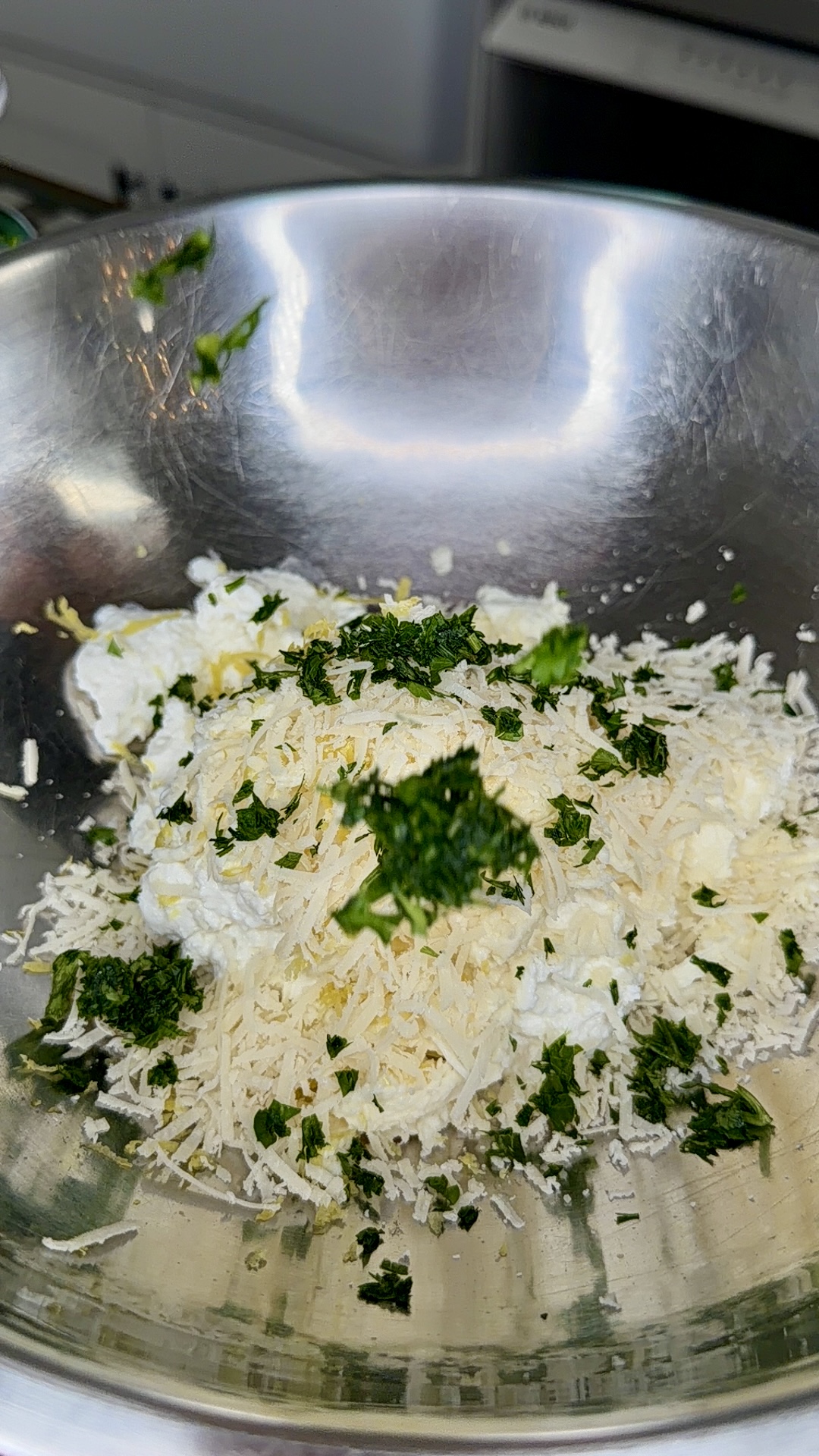 A stainless steel mixing bowl containing shredded cheese, ricotta cheese, and chopped fresh herbs, ready to be mixed together.
