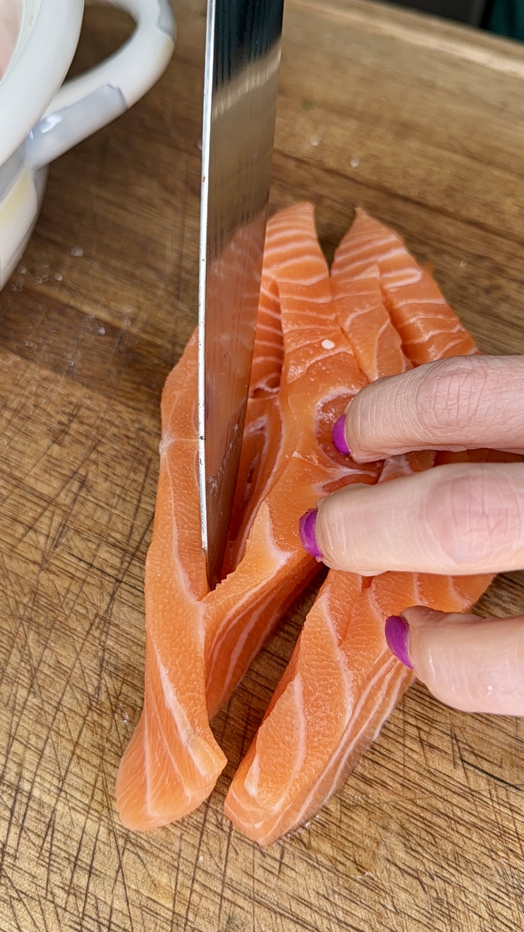 A hand with purple-painted nails holds raw salmon while a large knife slices it into thin strips on a wooden cutting board.