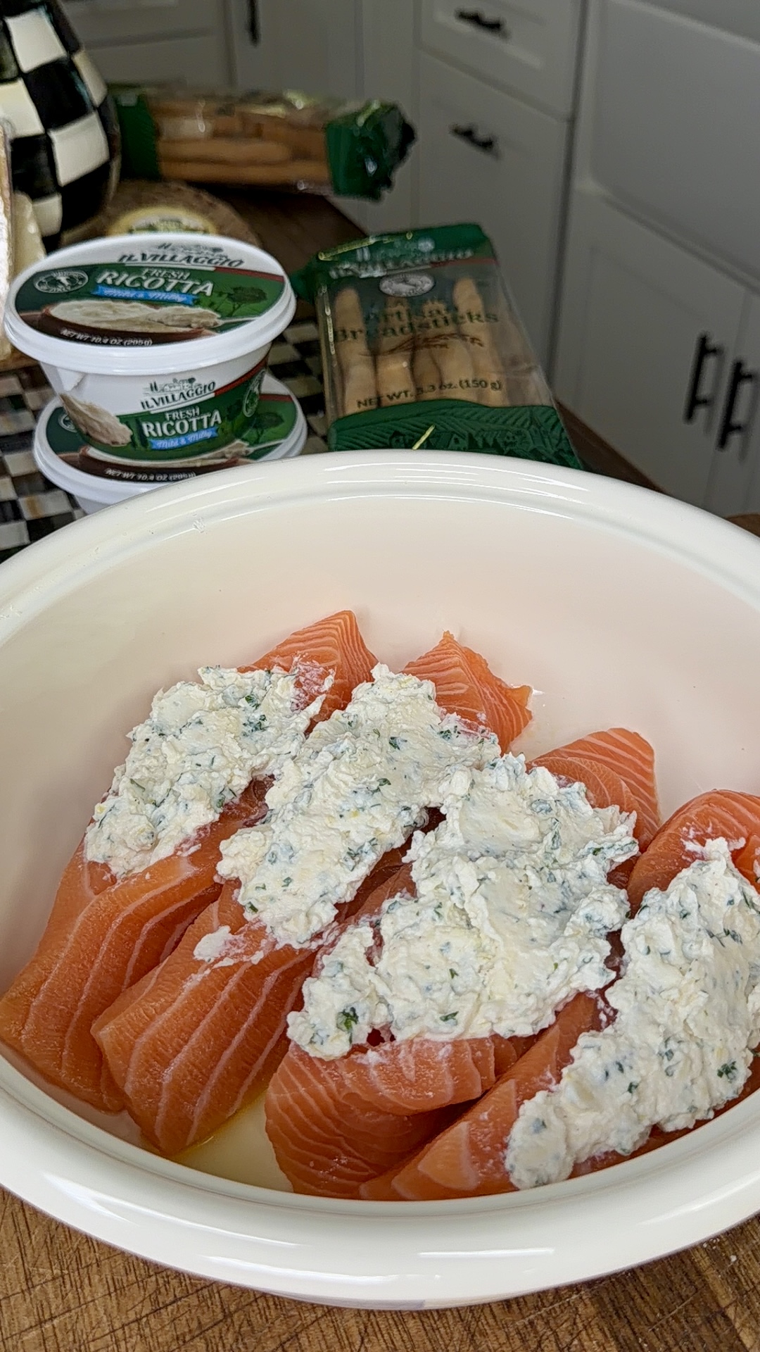 A white bowl with raw salmon fillets split open and stuffed with a creamy ricotta and herb filling. Containers of ricotta cheese and packages of cannelloni pasta are on the counter in the background.