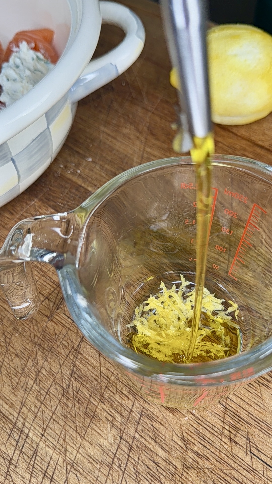 Olive oil being poured into a glass measuring cup containing lemon zest, on a wooden cutting board. A lemon and a bowl with cheese and tomato are visible in the background.