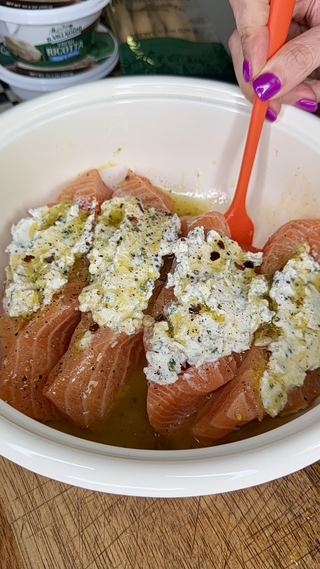 Four raw salmon fillets in a white bowl are topped with a creamy herb mixture and oil. A hand with purple nail polish holds an orange spoon next to the salmon.