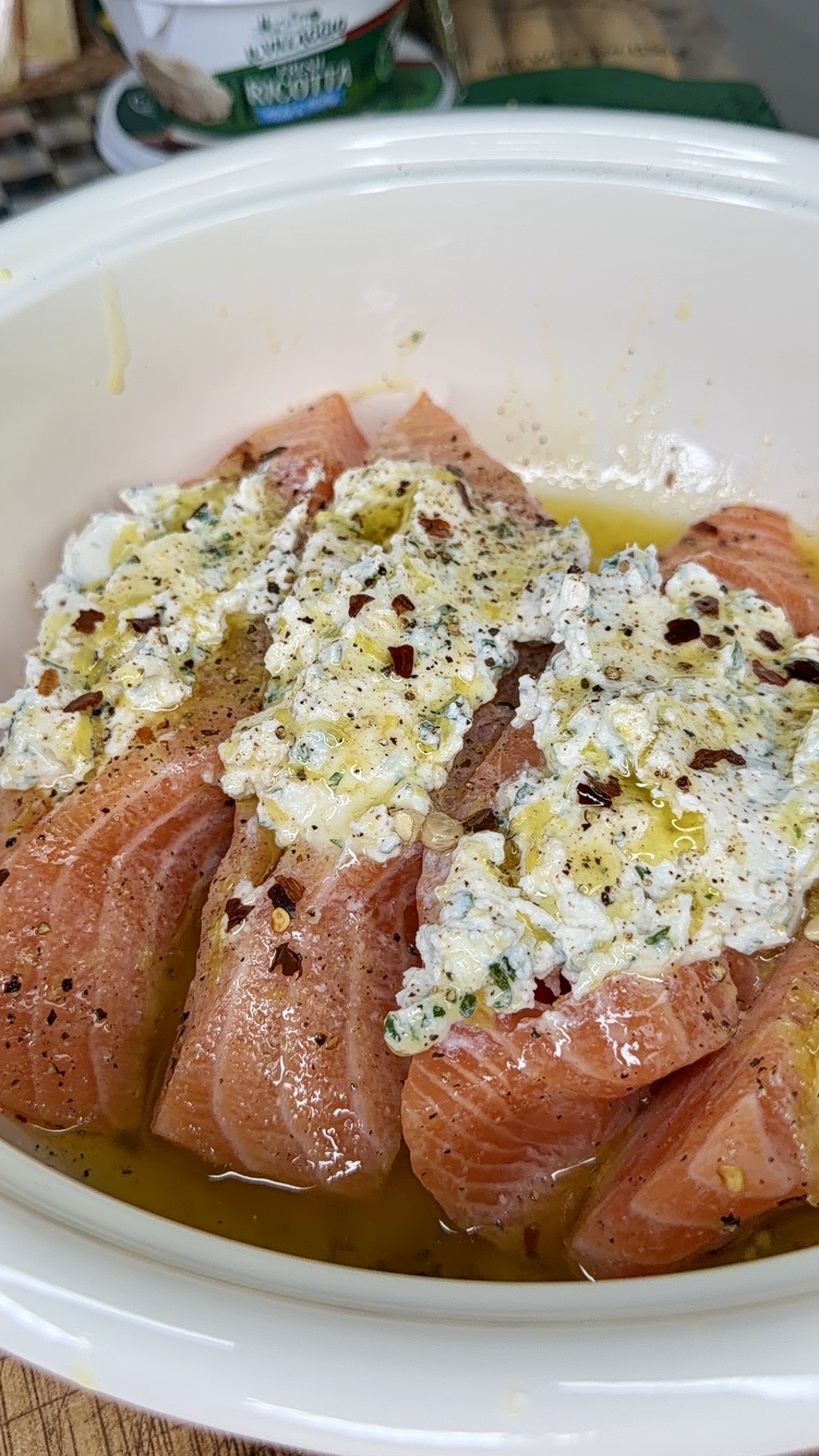 Raw salmon fillets in a white dish topped with a creamy herb mixture, black pepper, red pepper flakes, and drizzled with olive oil, ready to be cooked.