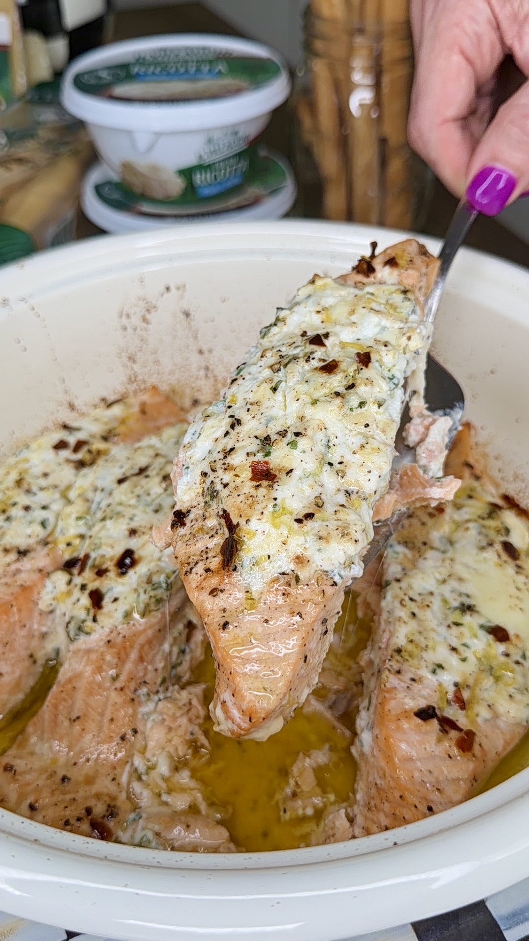 A hand with purple nail polish lifts a piece of baked salmon topped with creamy herb and cheese mixture from a dish, with more salmon pieces and melted butter visible below.