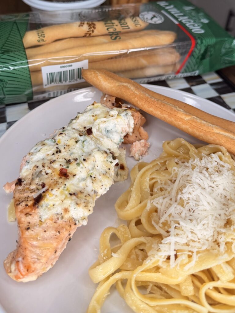 A plate with baked salmon topped with creamy cheese and herbs, fettuccine pasta sprinkled with grated cheese, and a breadstick. In the background, a package of Italian breadsticks is visible.
