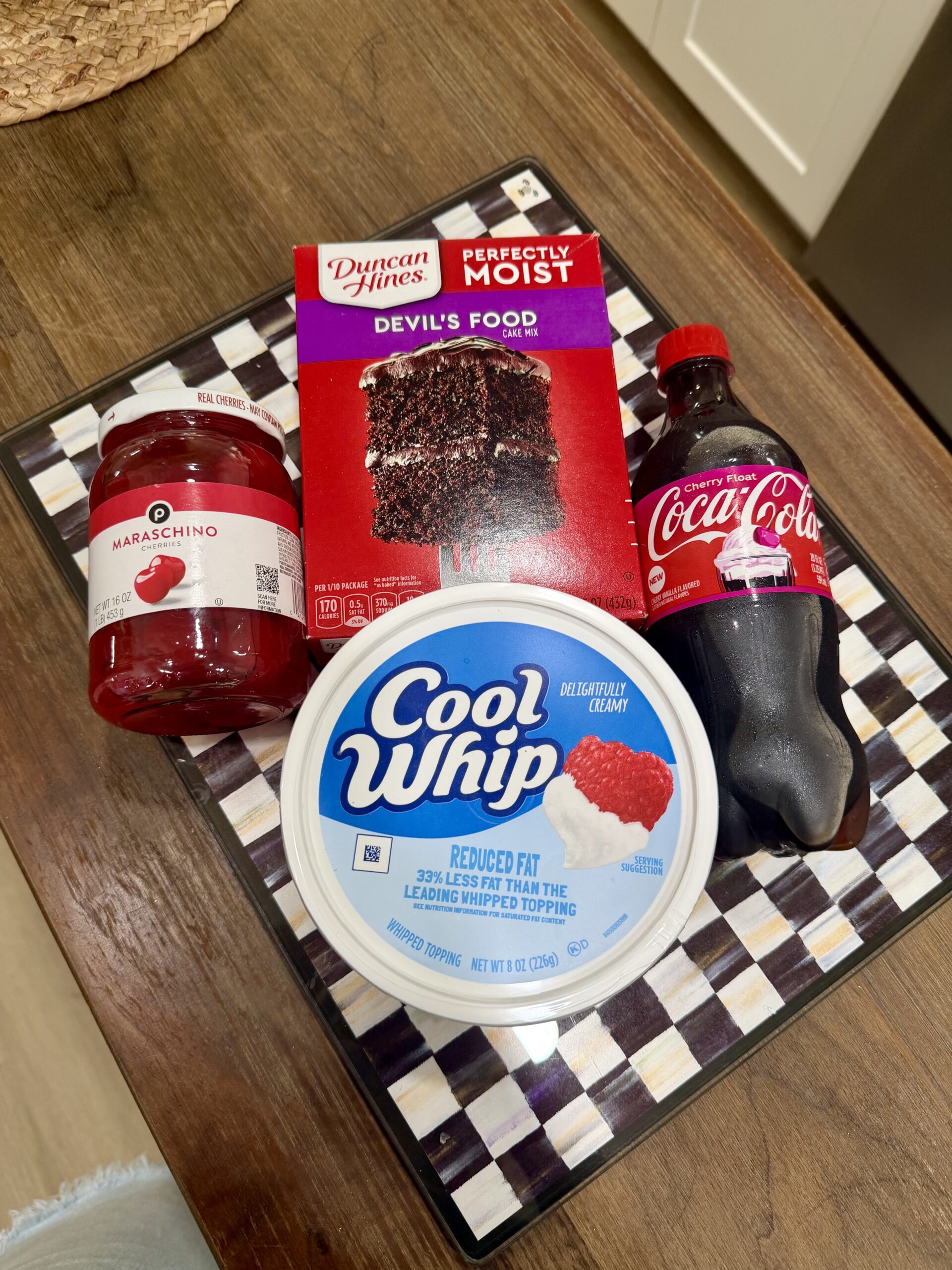 A jar of maraschino cherries, a box of Duncan Hines Devil’s Food cake mix, a bottle of Coca-Cola, and a tub of Cool Whip are arranged on a black and white checkered tray on a kitchen counter.