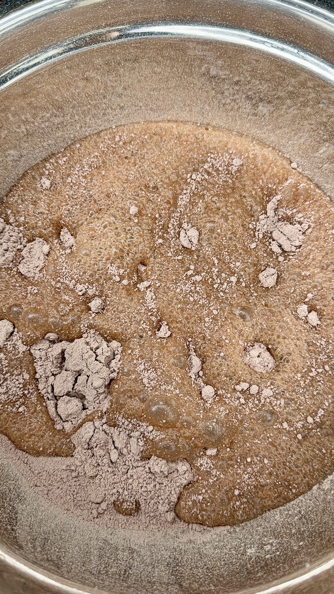 A metal mixing bowl containing a bubbly brown liquid with clumps of dry cocoa powder or cake mix scattered on top, partially dissolved into the mixture.