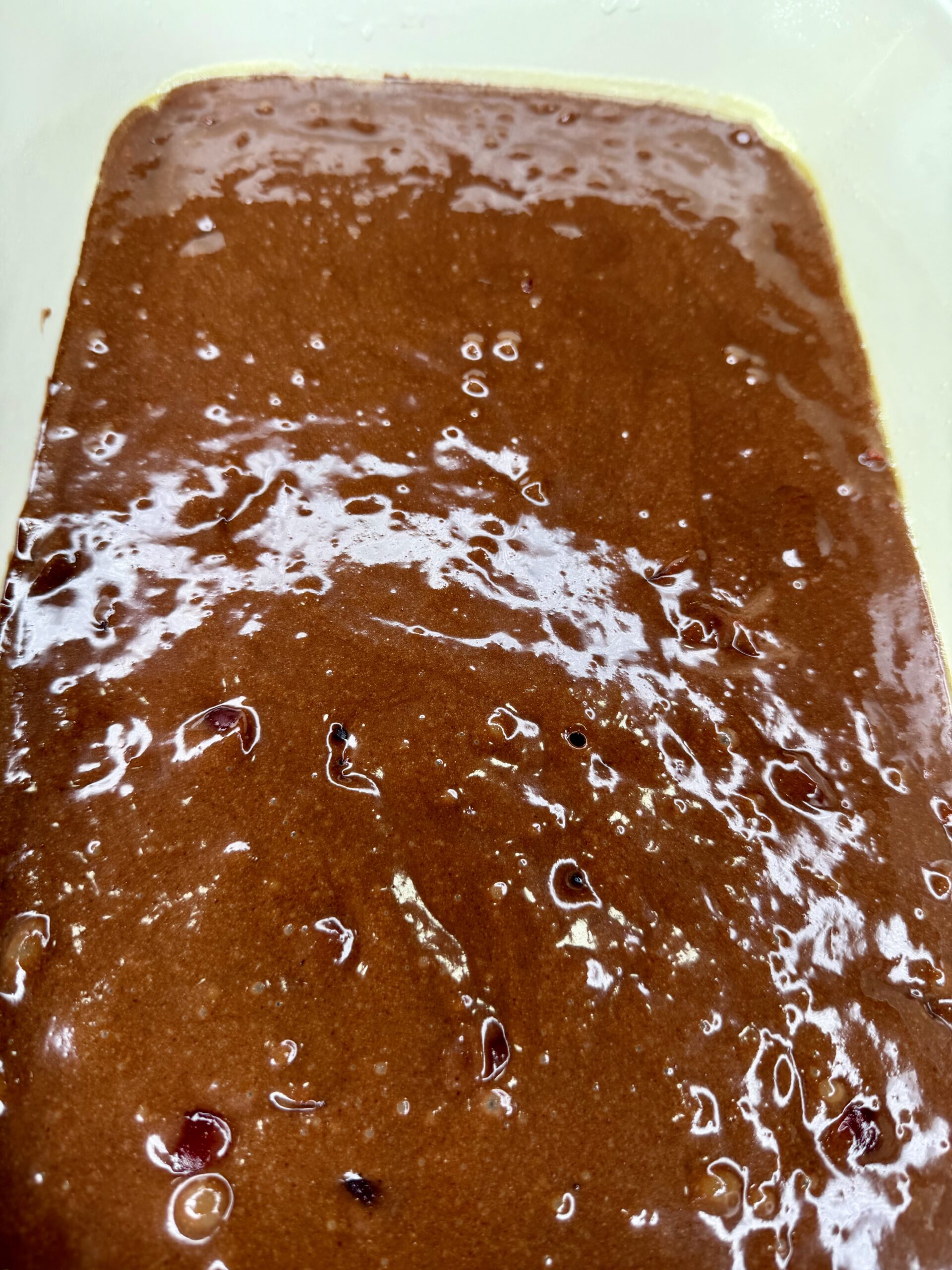 Close-up of chocolate brownie batter in a mixing bowl, showing a smooth, glossy texture with small air bubbles and bits of chocolate or other ingredients visible throughout.