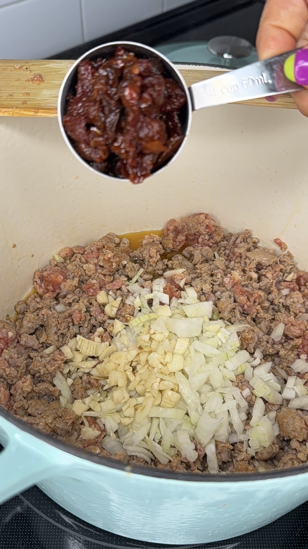 Ground beef is browning in a pot with chopped onions and garlic on top, while a hand holds a measuring cup filled with chopped sun-dried tomatoes above the pot, ready to add them.